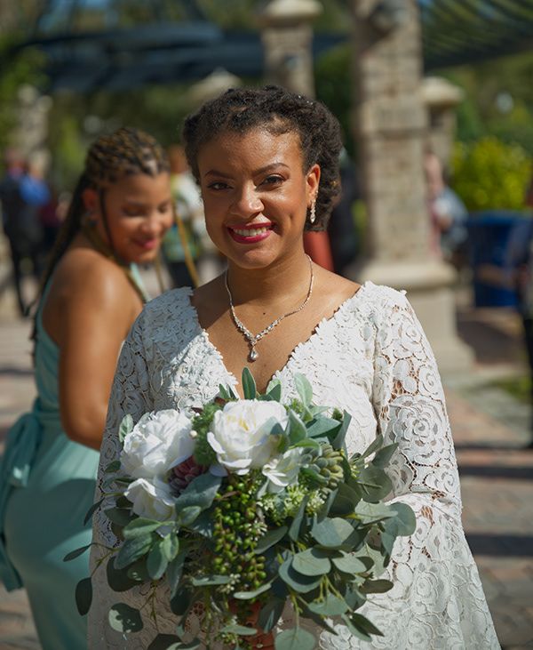 A bride in a white lace dress holds a bouquet with white roses and eucalyptus at an outdoor wedding.