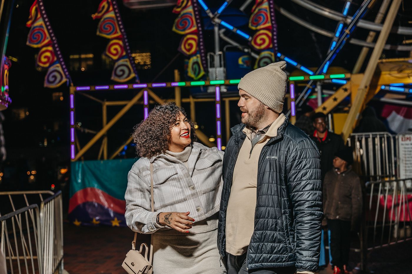 A couple converses at a nighttime carnival with colorful lights in the background.