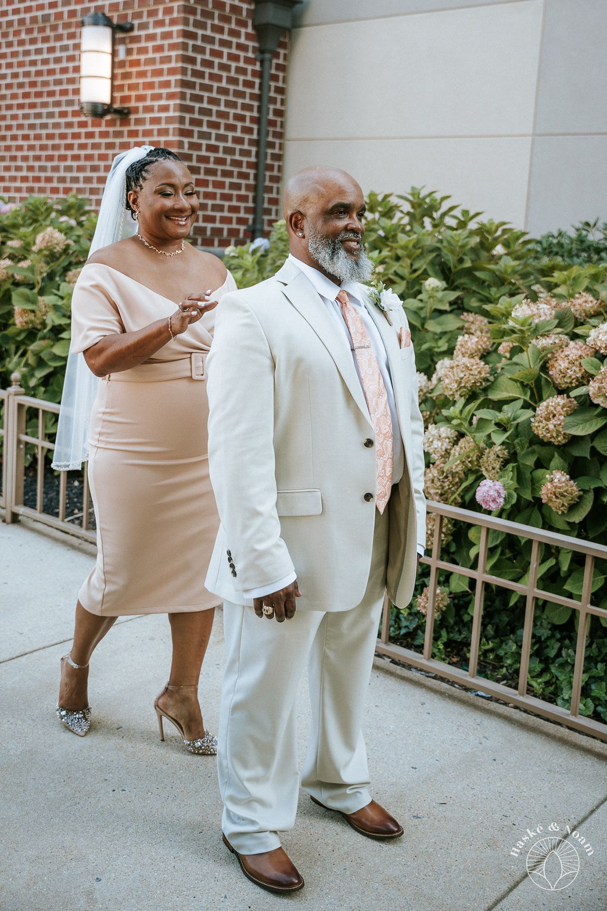 A couple in formal attire posing outside a brick building with blooming hydrangeas.