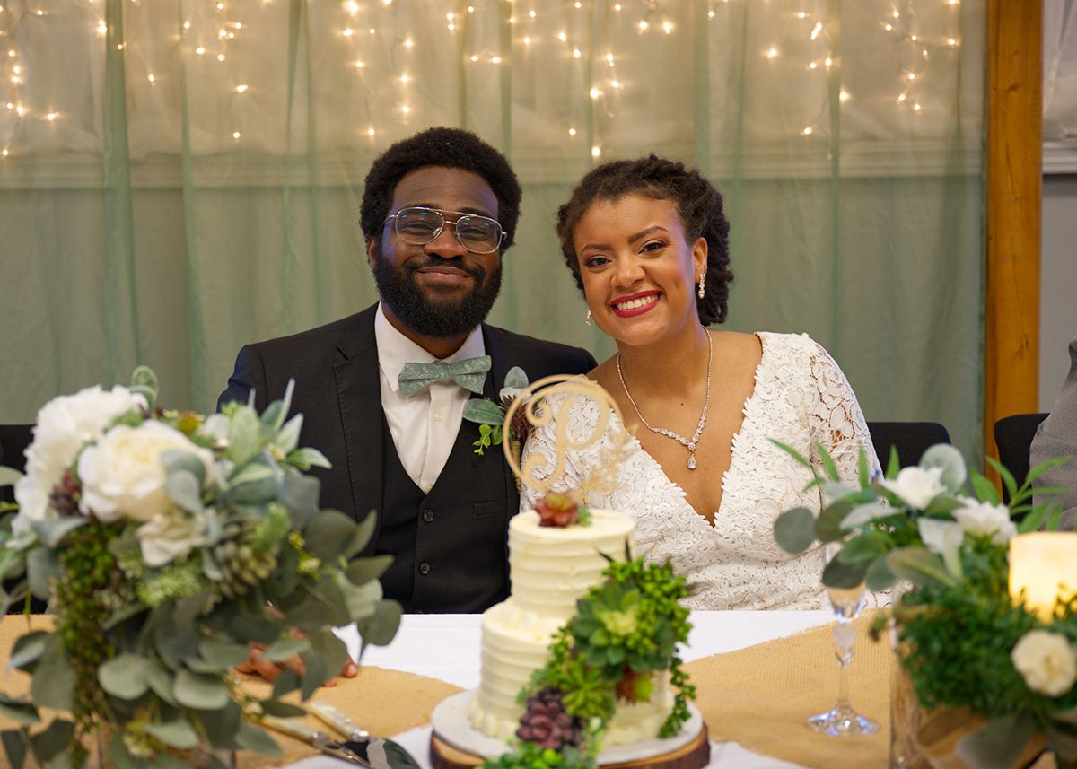Happy couple sitting at reception table with wedding cake and floral decorations.