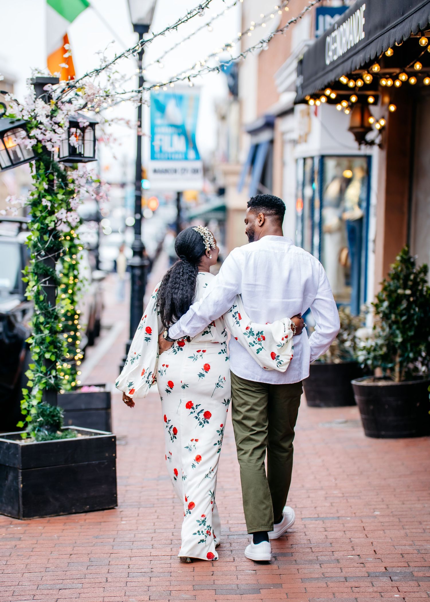 A couple wearing matching floral outfits walks together on a brick street decorated with holiday lights and greenery.