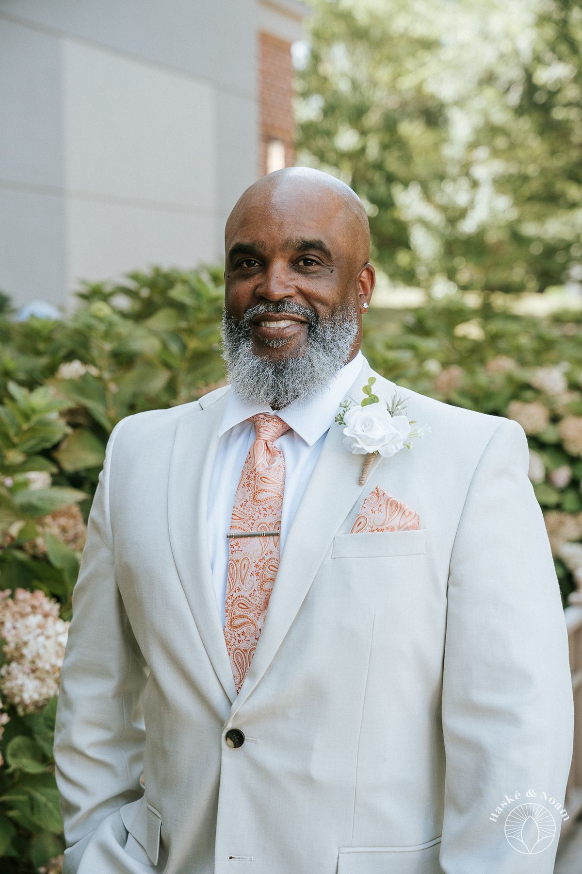 Elegant groom in white suit with peach tie and pocket square standing outdoors.