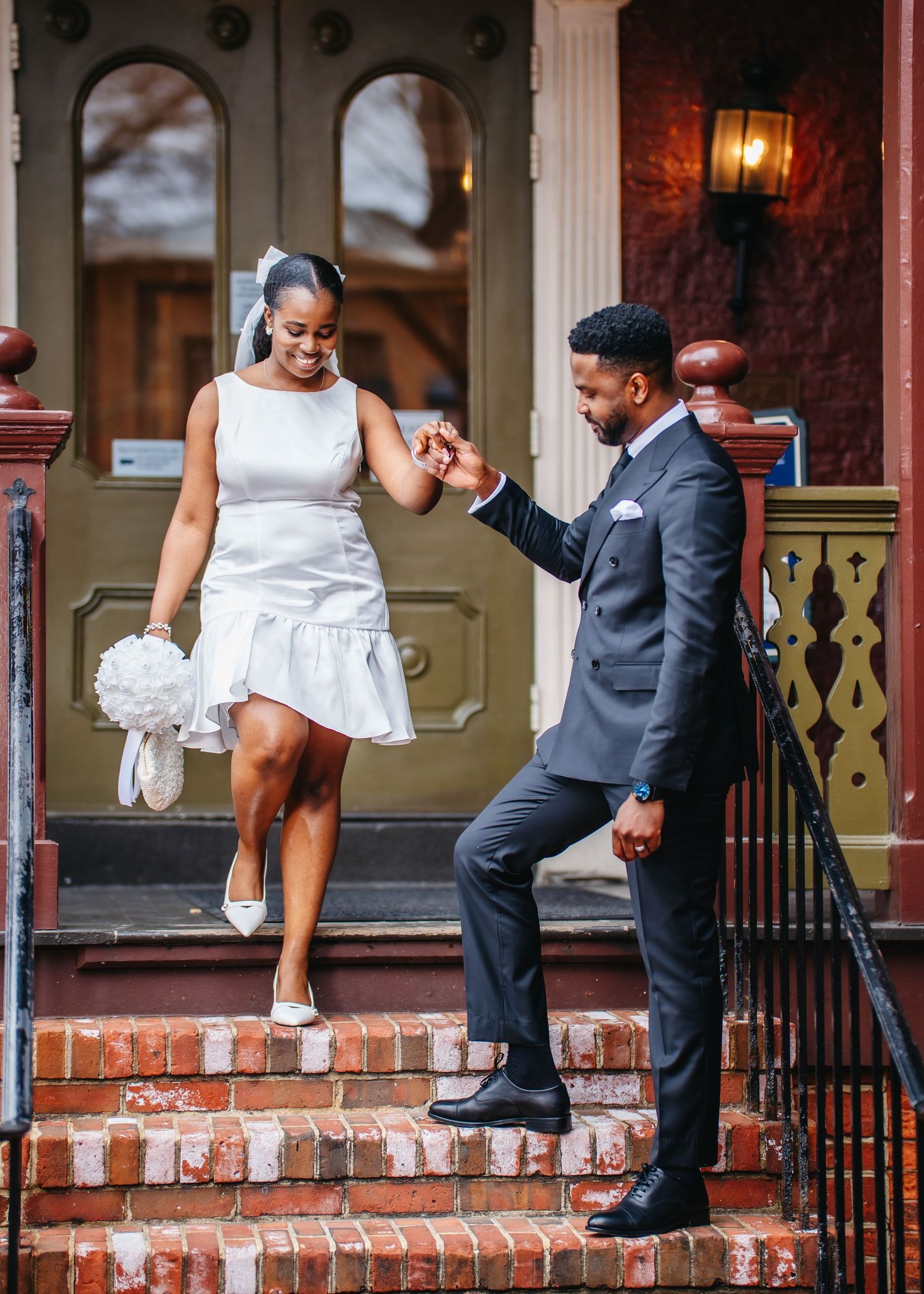 A couple on brick steps share a joyful moment, the bride in a short white dress holding a bouquet and groom in a grey suit.