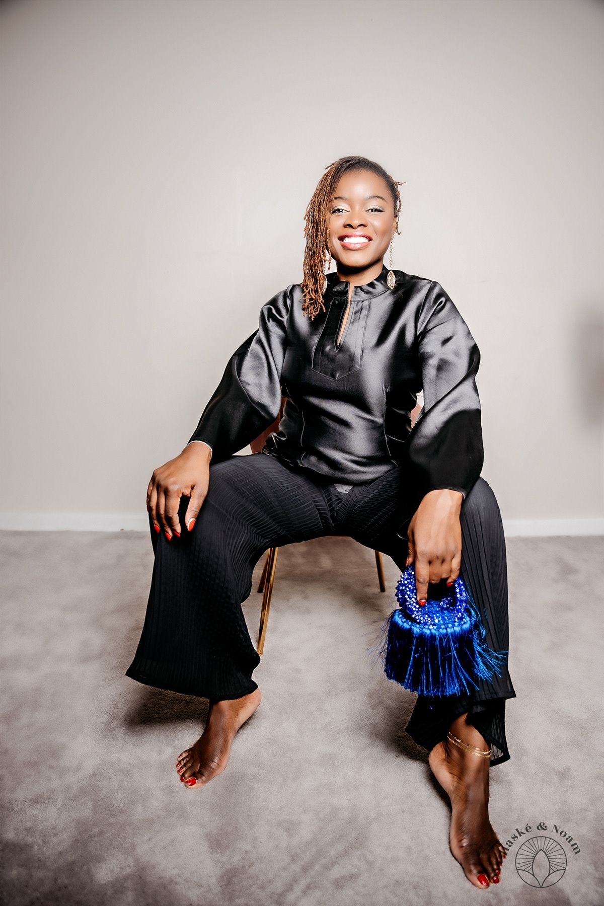 The person in a flowing black silk outfit sits casually barefoot on a gray floor, holding a blue beaded handbag.