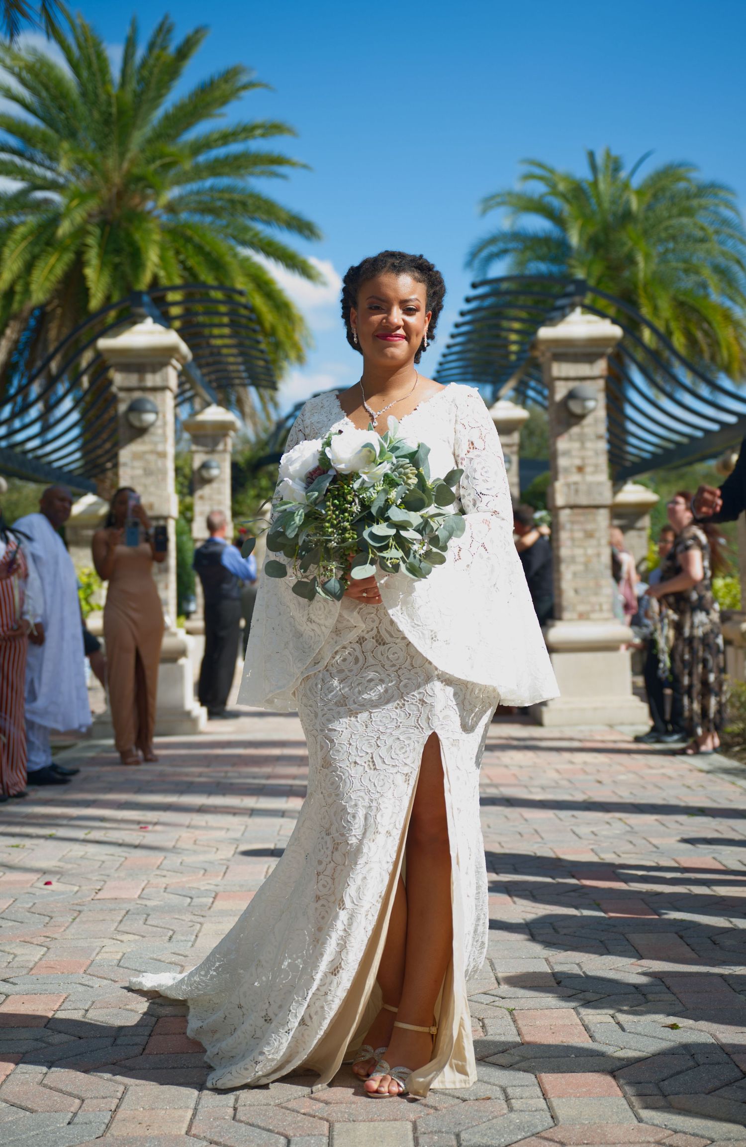 Bride in white lace gown with bell sleeves holds bouquet in front of palm trees and stone pillars.