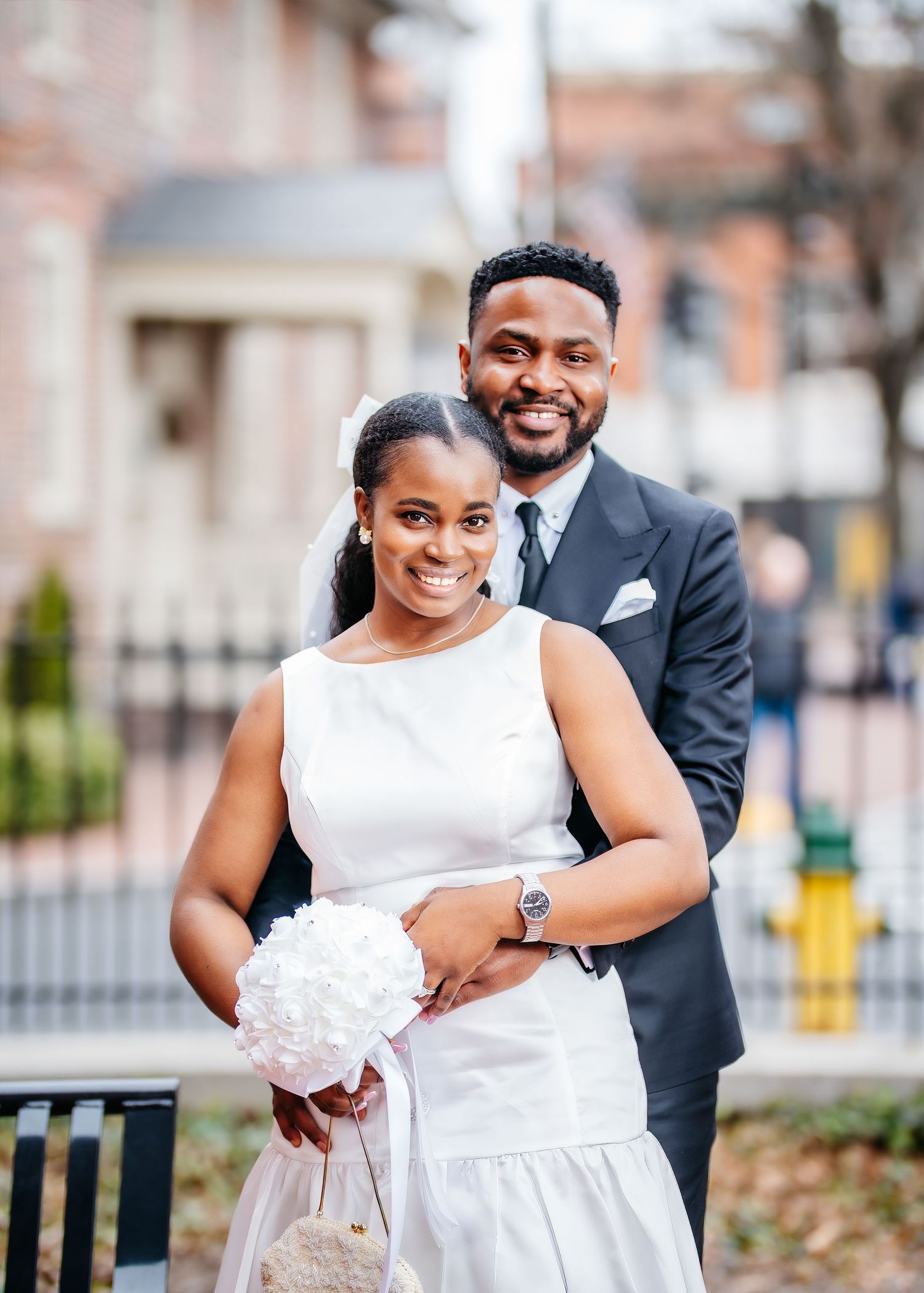 A couple dressed in formal wedding attire embrace outdoors while holding a white bouquet.