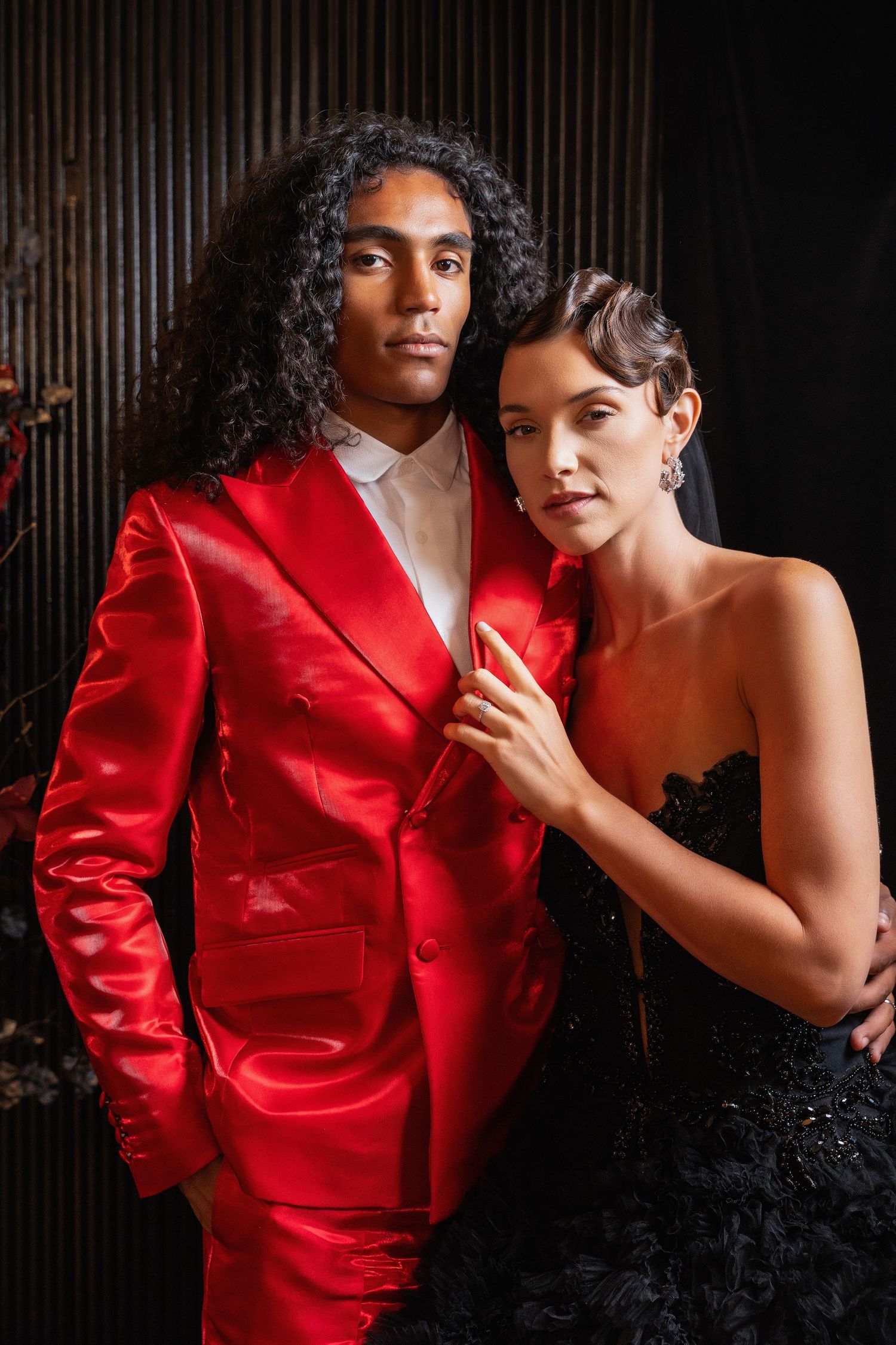 Glamorous couple posing in red silk suit and black evening gown against textured wall.