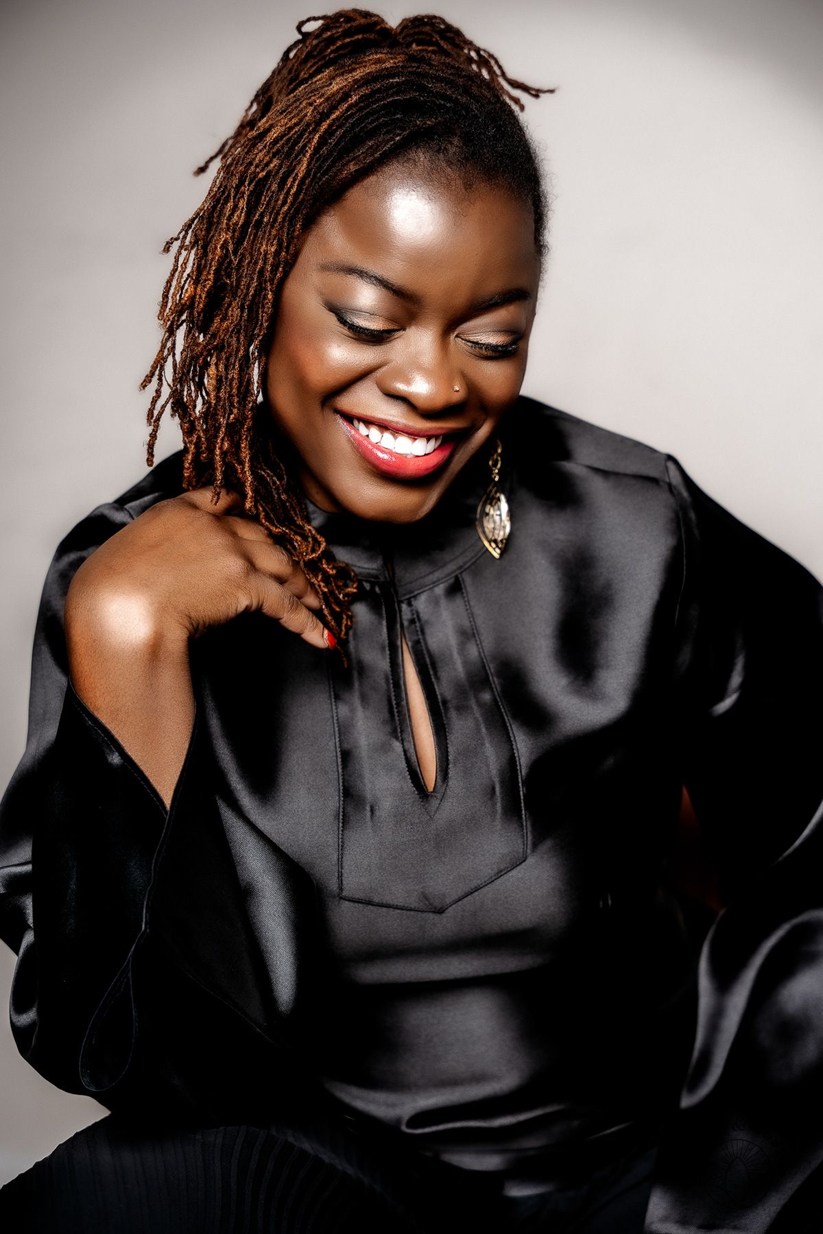 A warm, joyful smile radiates as locs frame the face, wearing an elegant black silk blouse with tie detail.