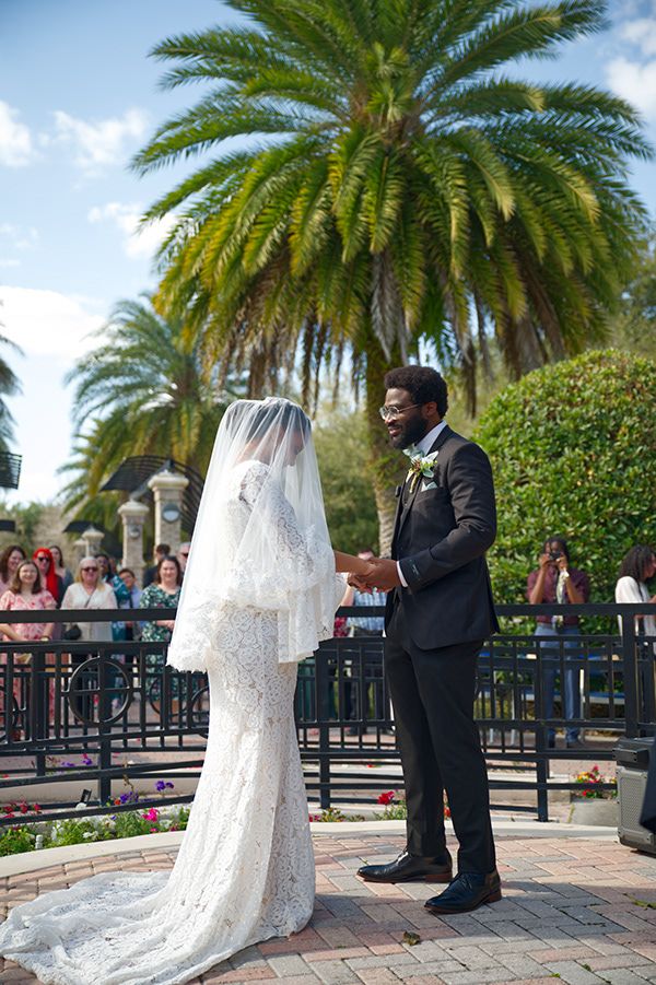 A wedding ceremony under palm trees with a bride in white lace gown and groom in dark suit.