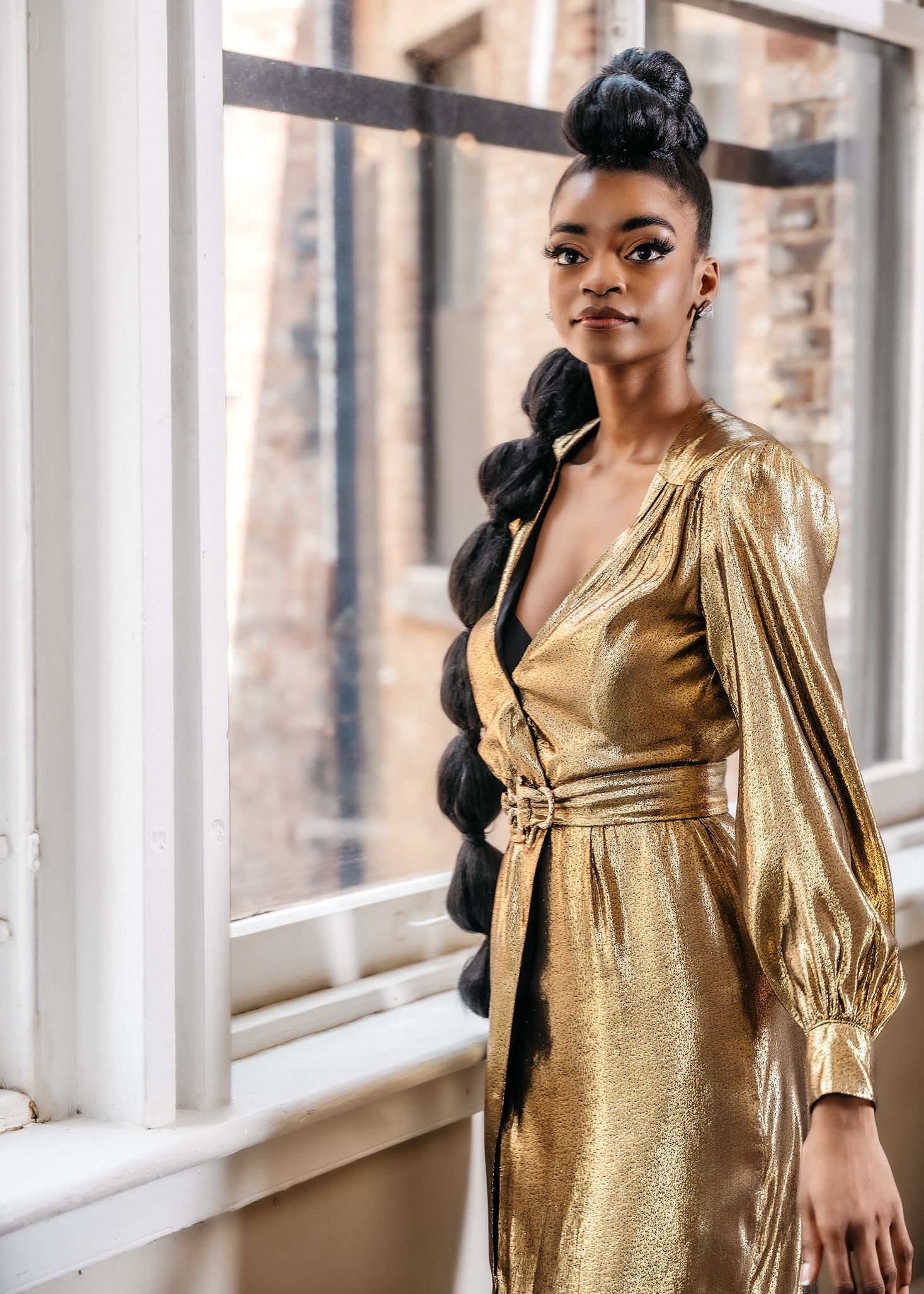 Model wearing a luxurious gold metallic wrap dress with balloon sleeves and bubble ponytail hairstyle near a window.