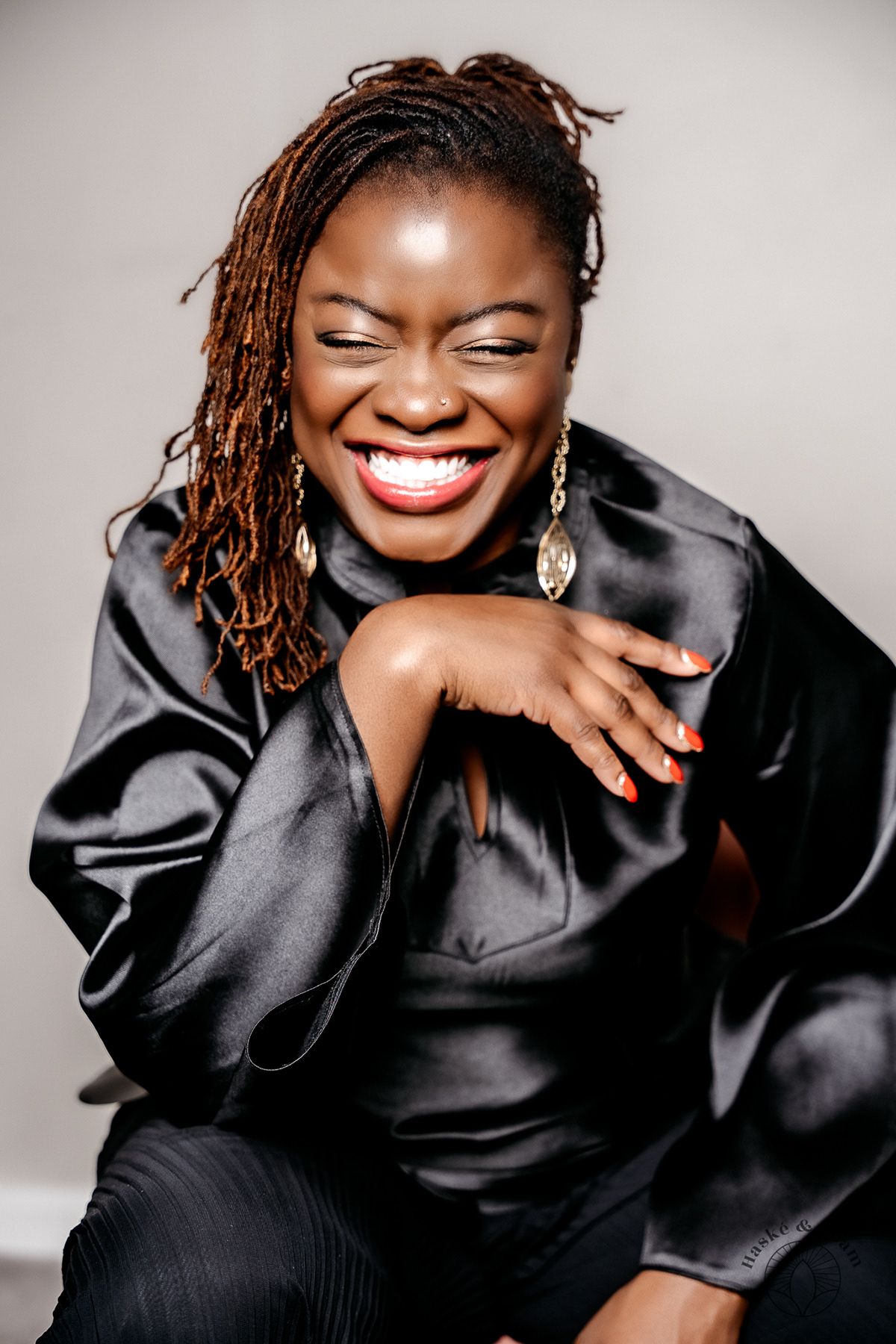 Professional portrait of someone in a black satin blouse laughing joyfully with dreadlocks and dangling earrings.