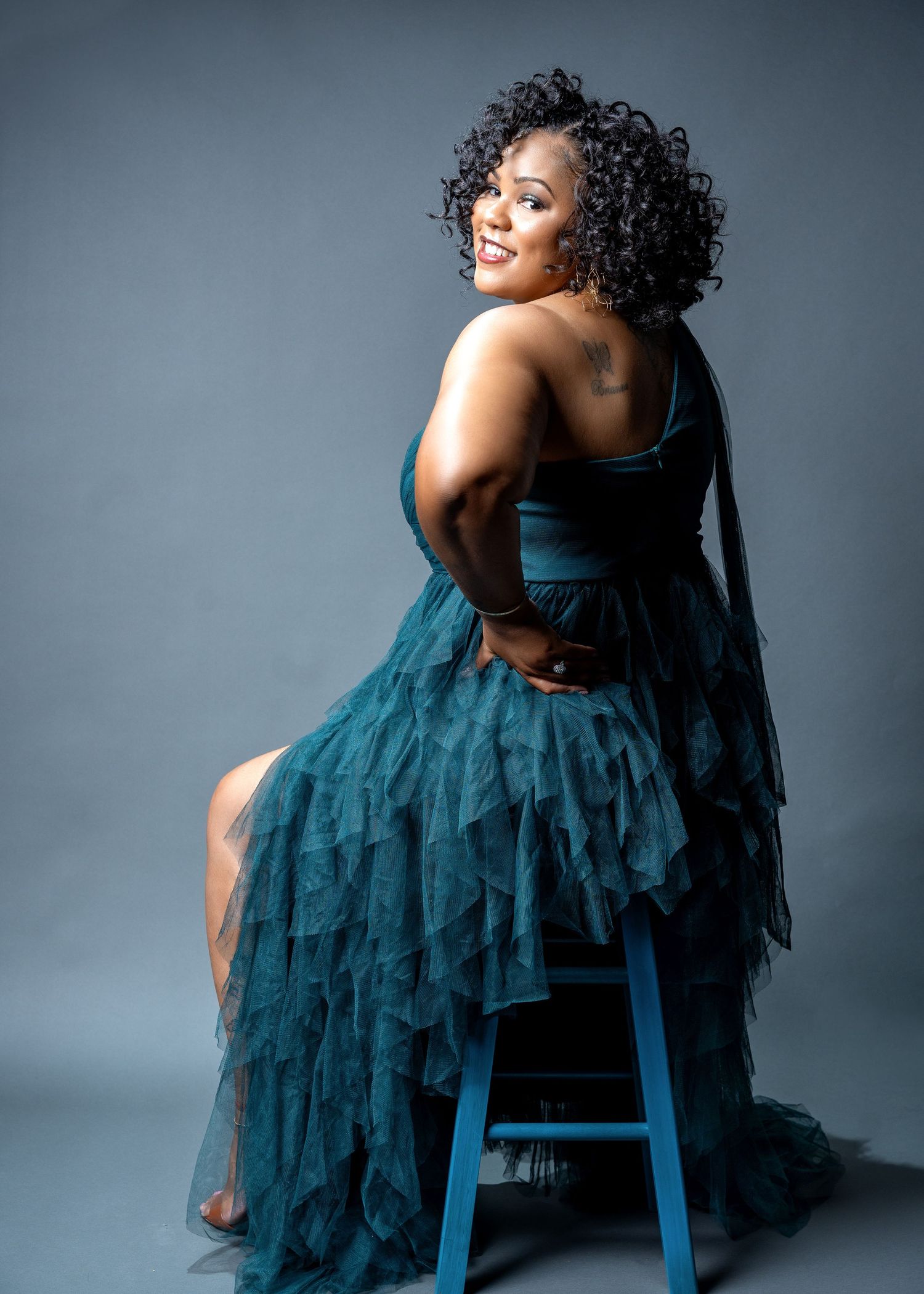 A glamorous portrait of someone in a teal evening gown posing on a blue stool against a gray background.