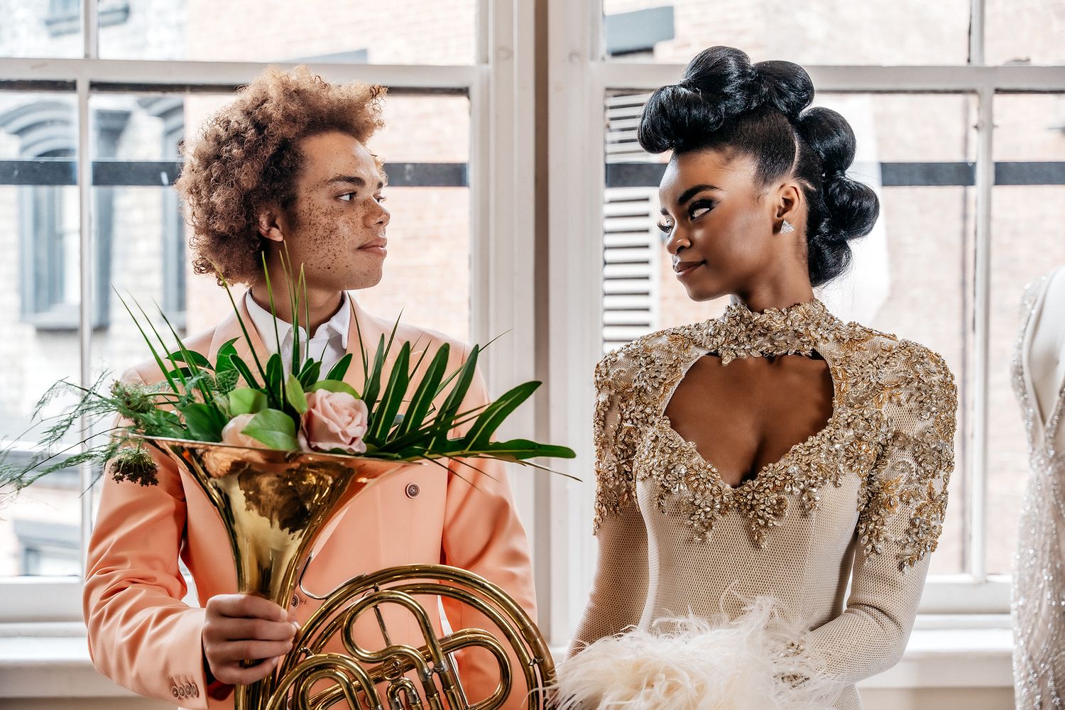 Fashion duo pose by window with tropical bouquet and elegant attire.