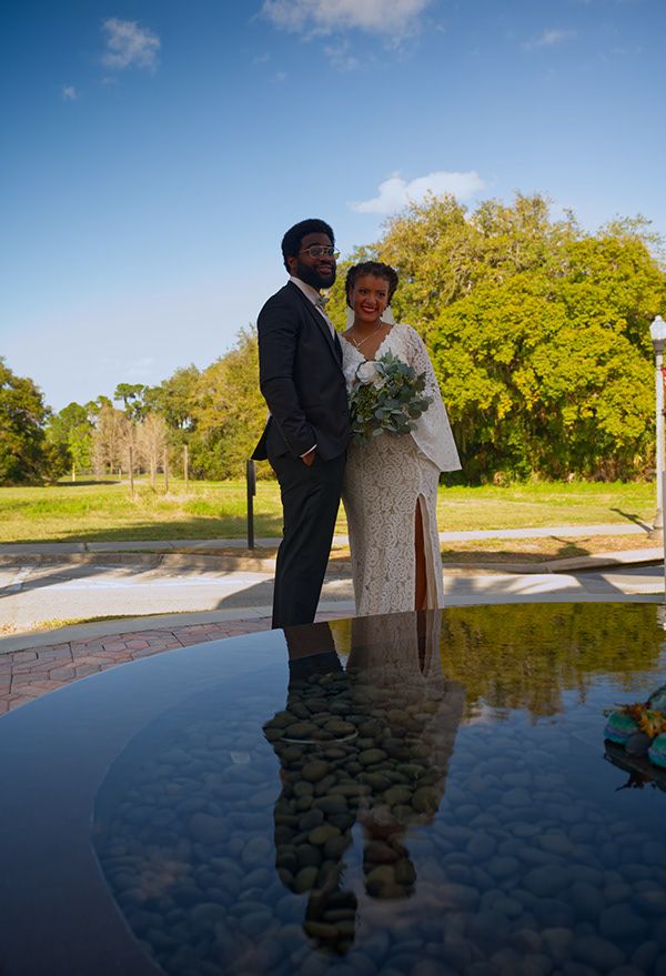 A couple in wedding attire stands reflected in a pool of water on their special day.