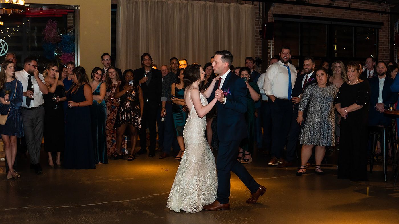 Couple shares their first dance at a wedding reception while guests watch from the edges of a dimly lit dance floor.