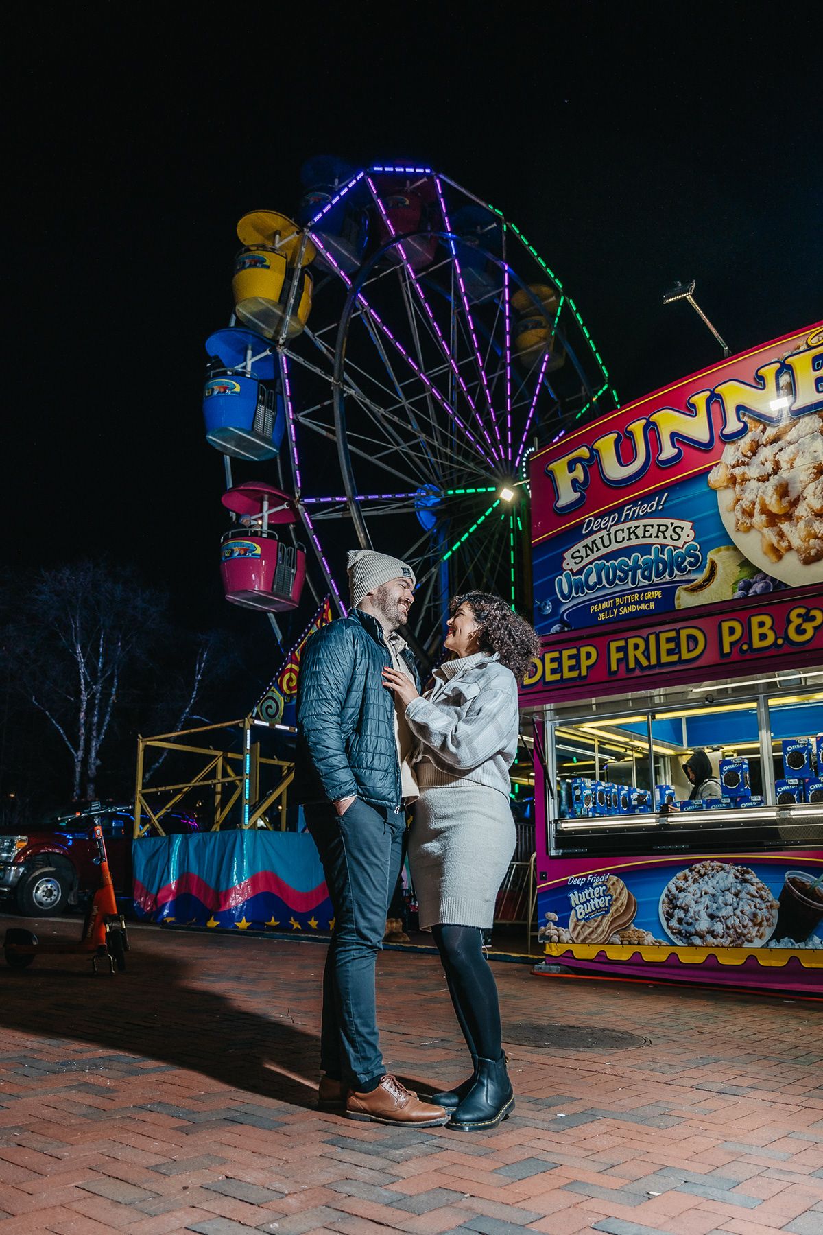 A couple embraces in front of an illuminated Ferris wheel and carnival food stand at night.