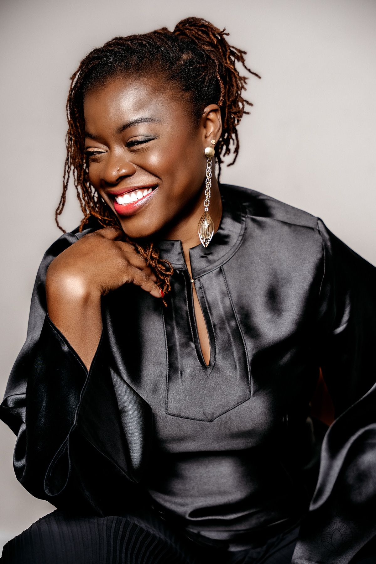 Portrait of someone wearing a black satin blouse while smiling joyfully against a light gray background.