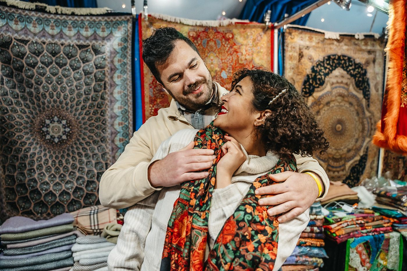 Couple laughing while shopping at a textile market with colorful tapestries displayed behind them.