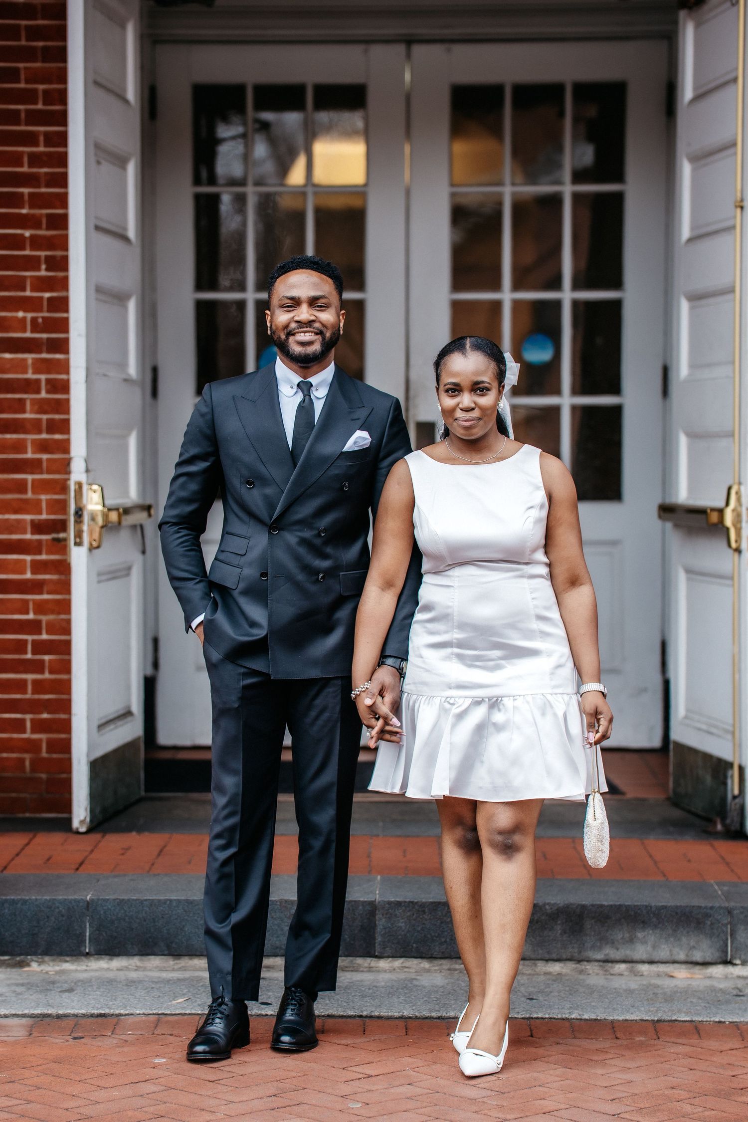 A couple stands together on brick steps outside white double doors, dressed formally in a navy suit and white dress.