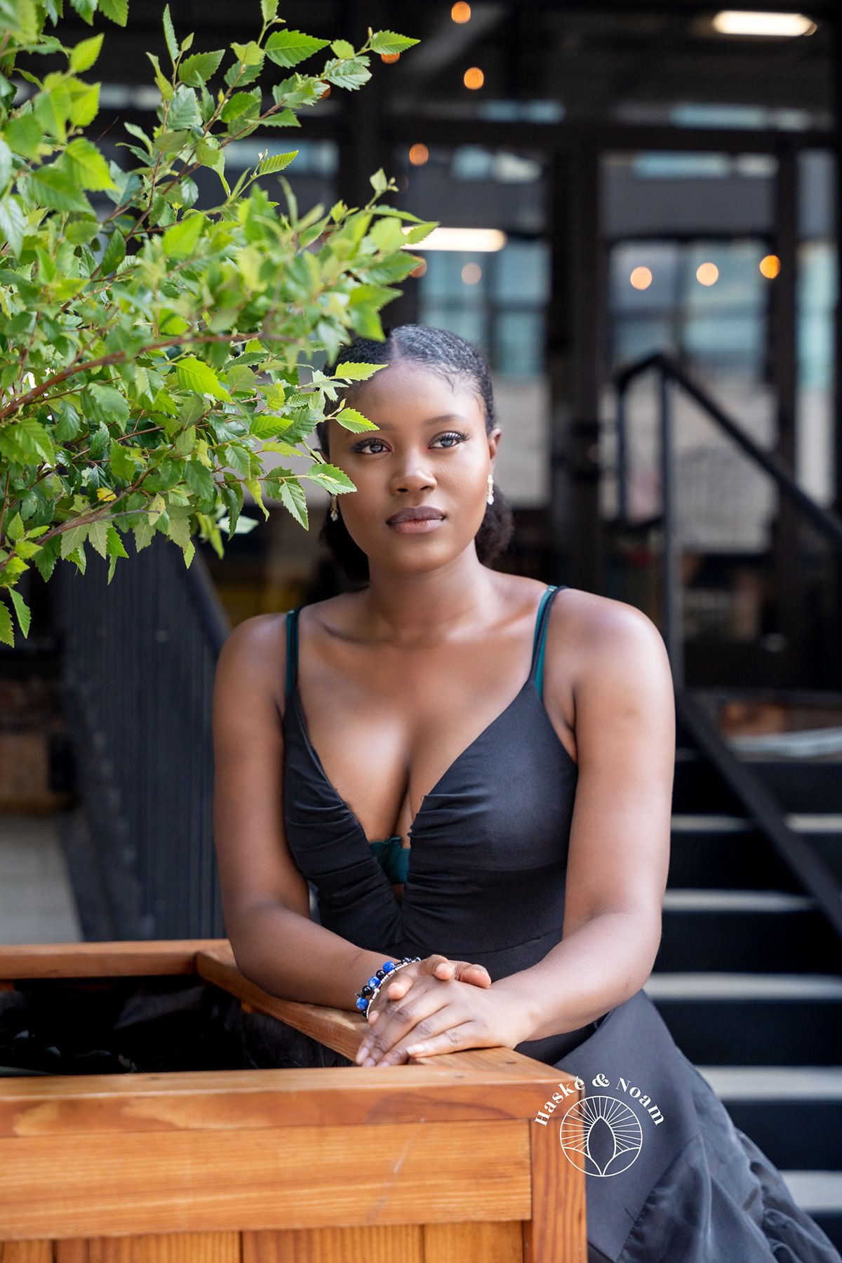 Portrait of someone in black dress seated at wooden table with greenery in background.