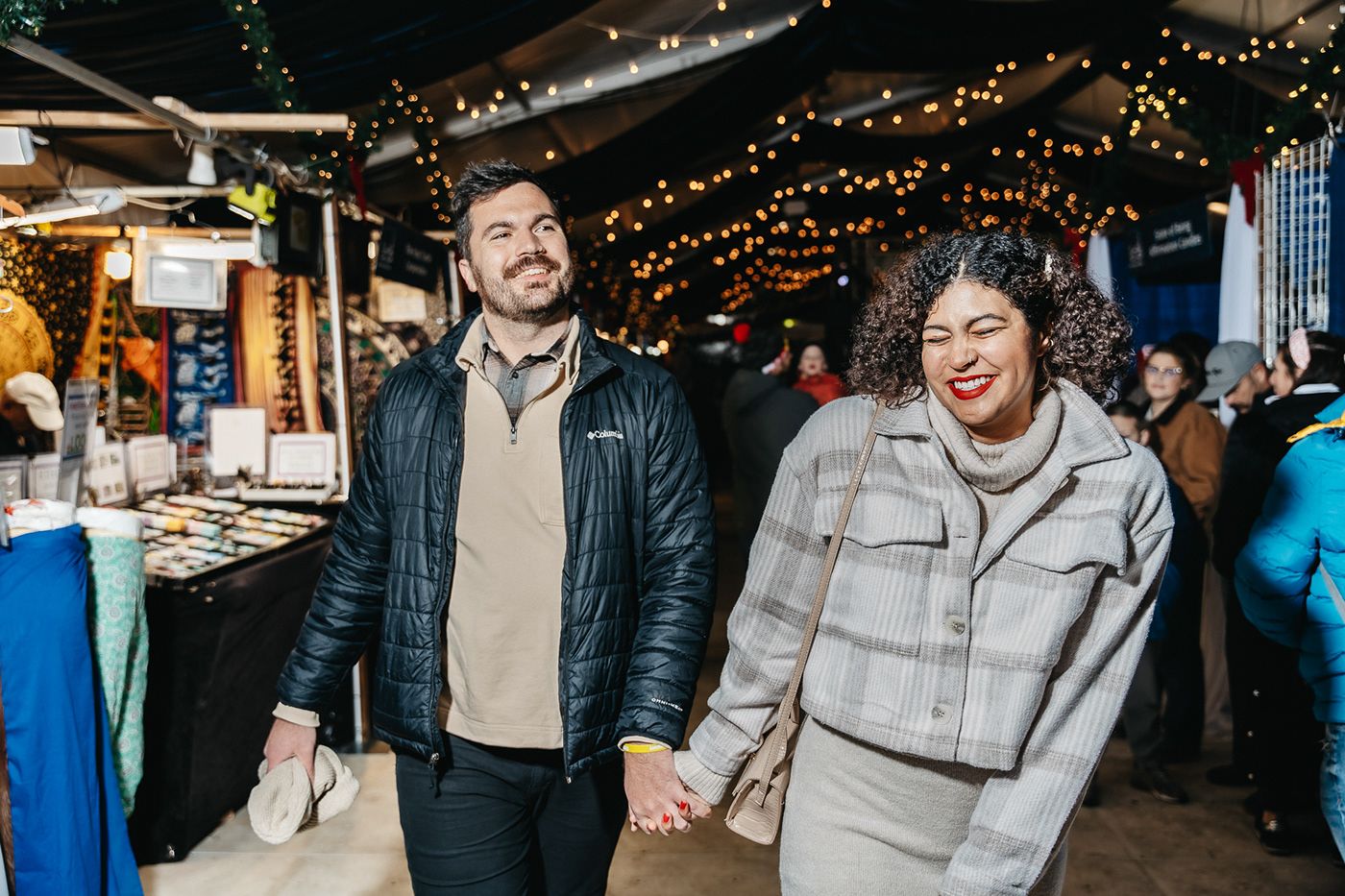 Two people laughing together while holding hands at an outdoor market with string lights in the background.