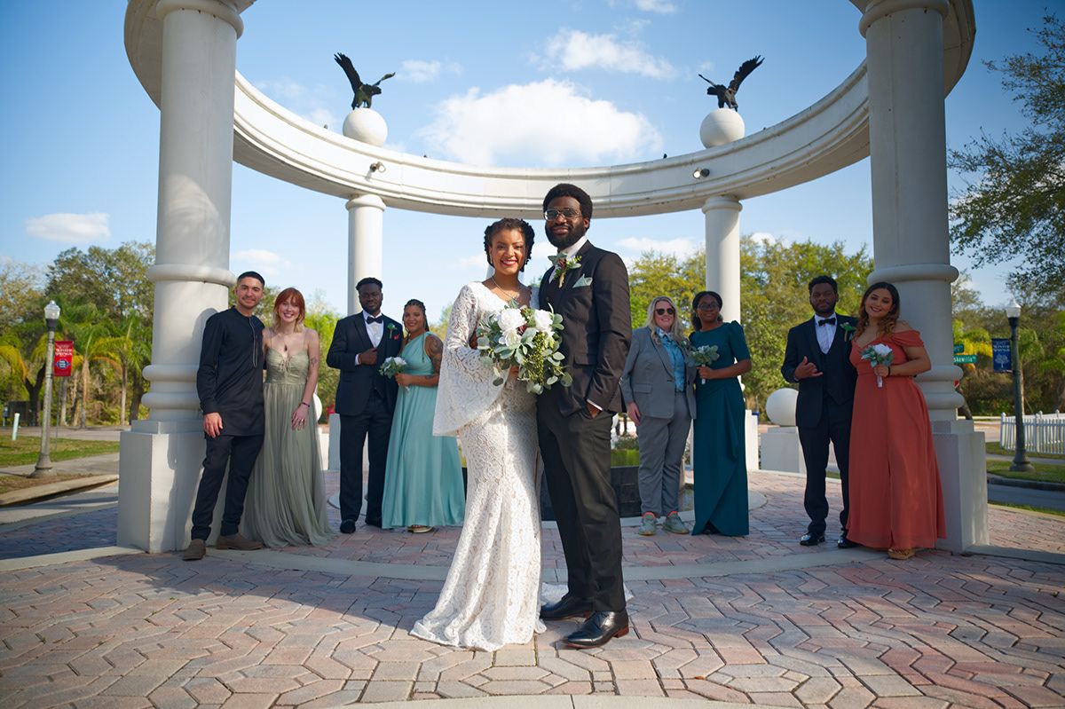 A wedding party poses under a white columned arch with blue skies in the background.