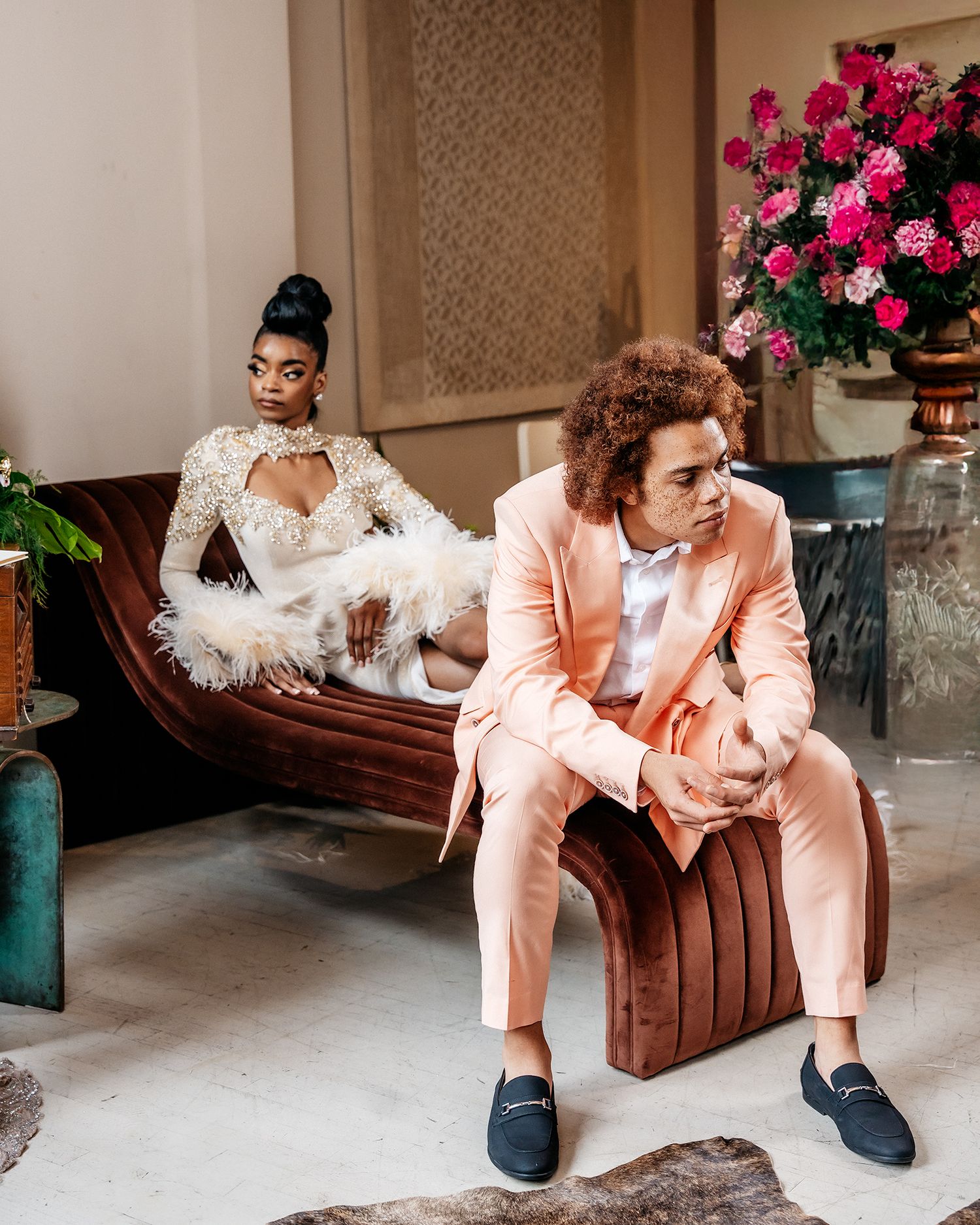 Fashion scene with white feathered gown and person in peach suit on vintage brown leather couch.