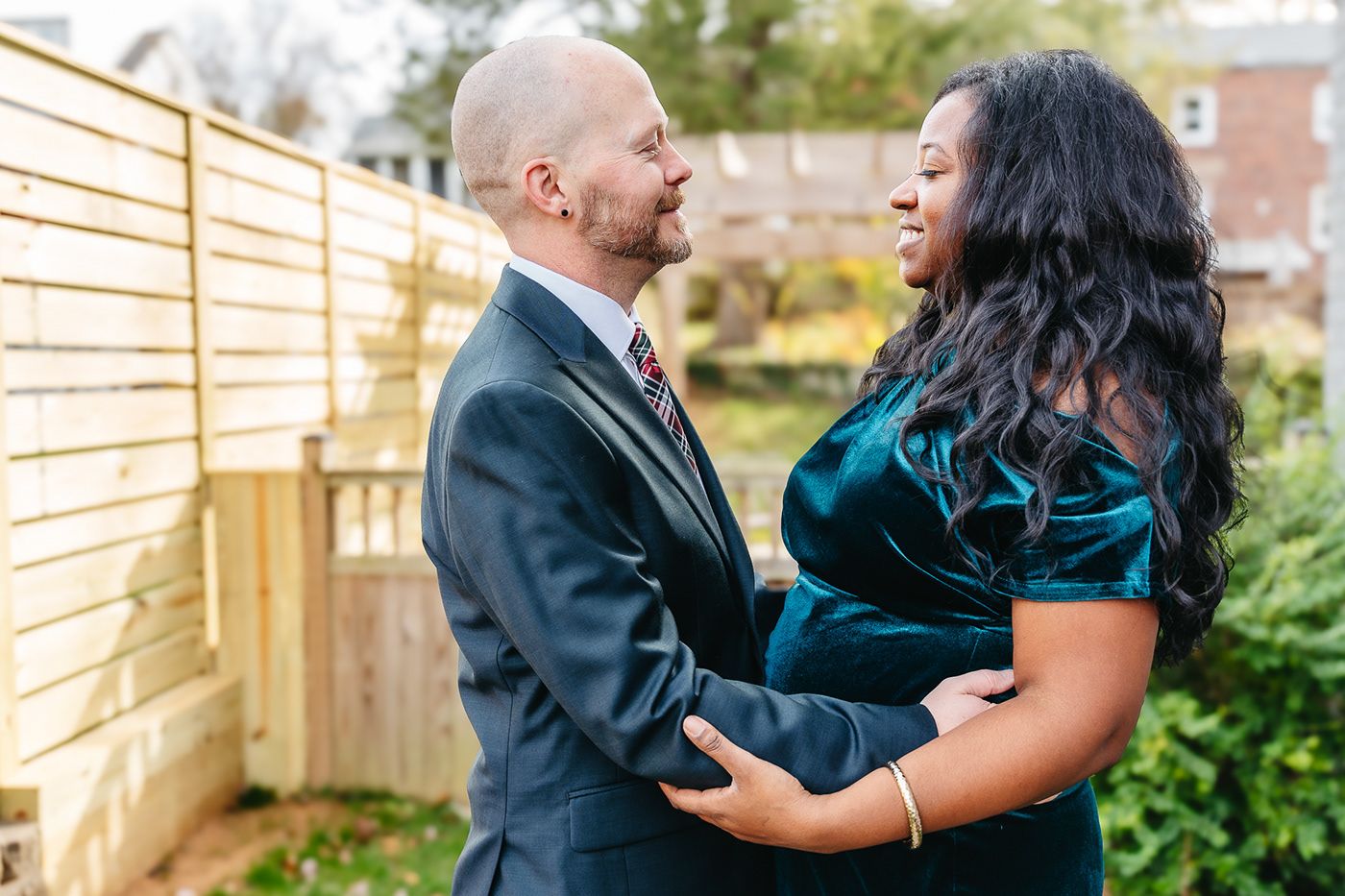 A couple embraces outdoors in front of a wooden fence, wearing formal attire in contrasting colors.