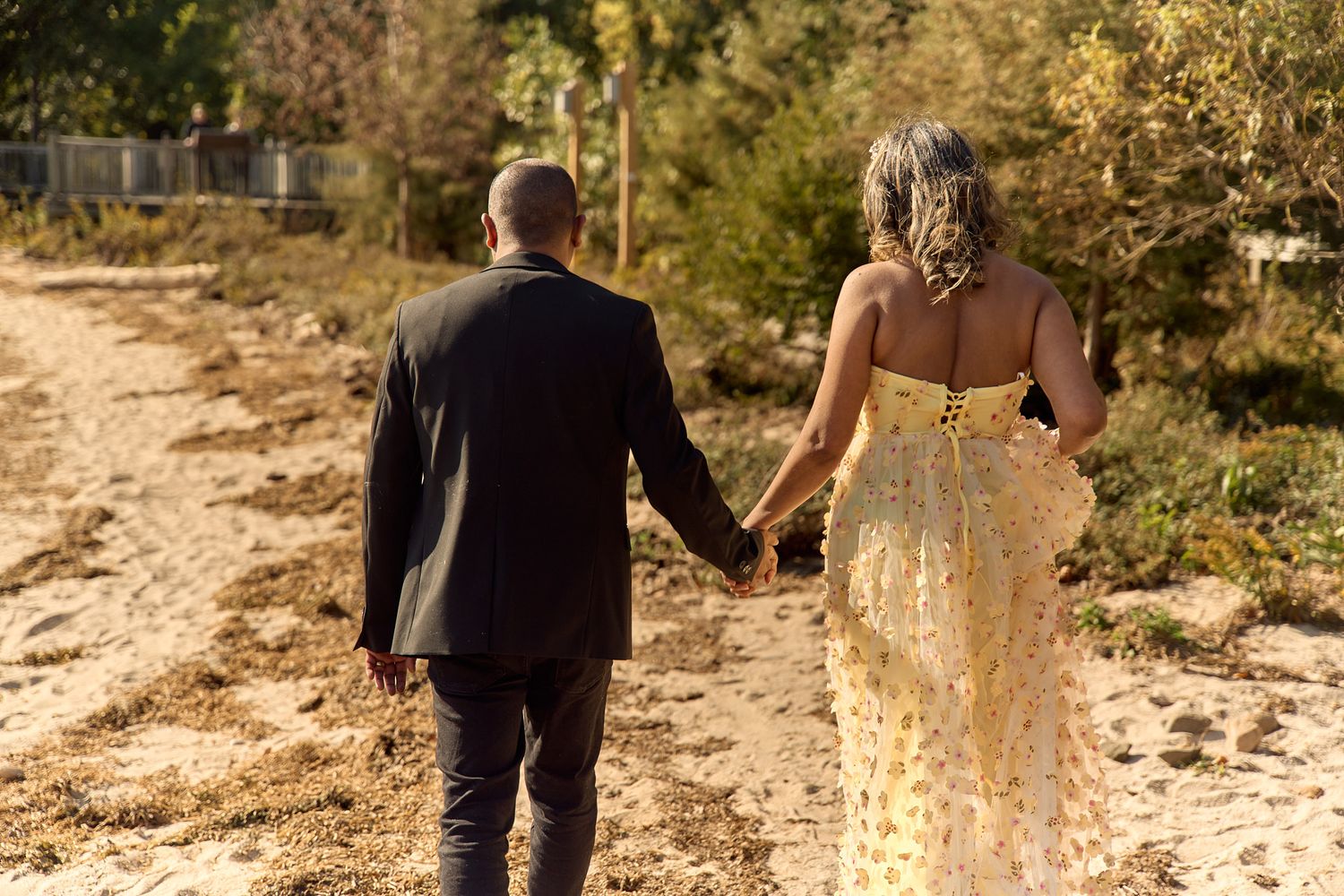A couple walking hand in hand on a dirt path wearing formal attire in golden autumn sunlight.