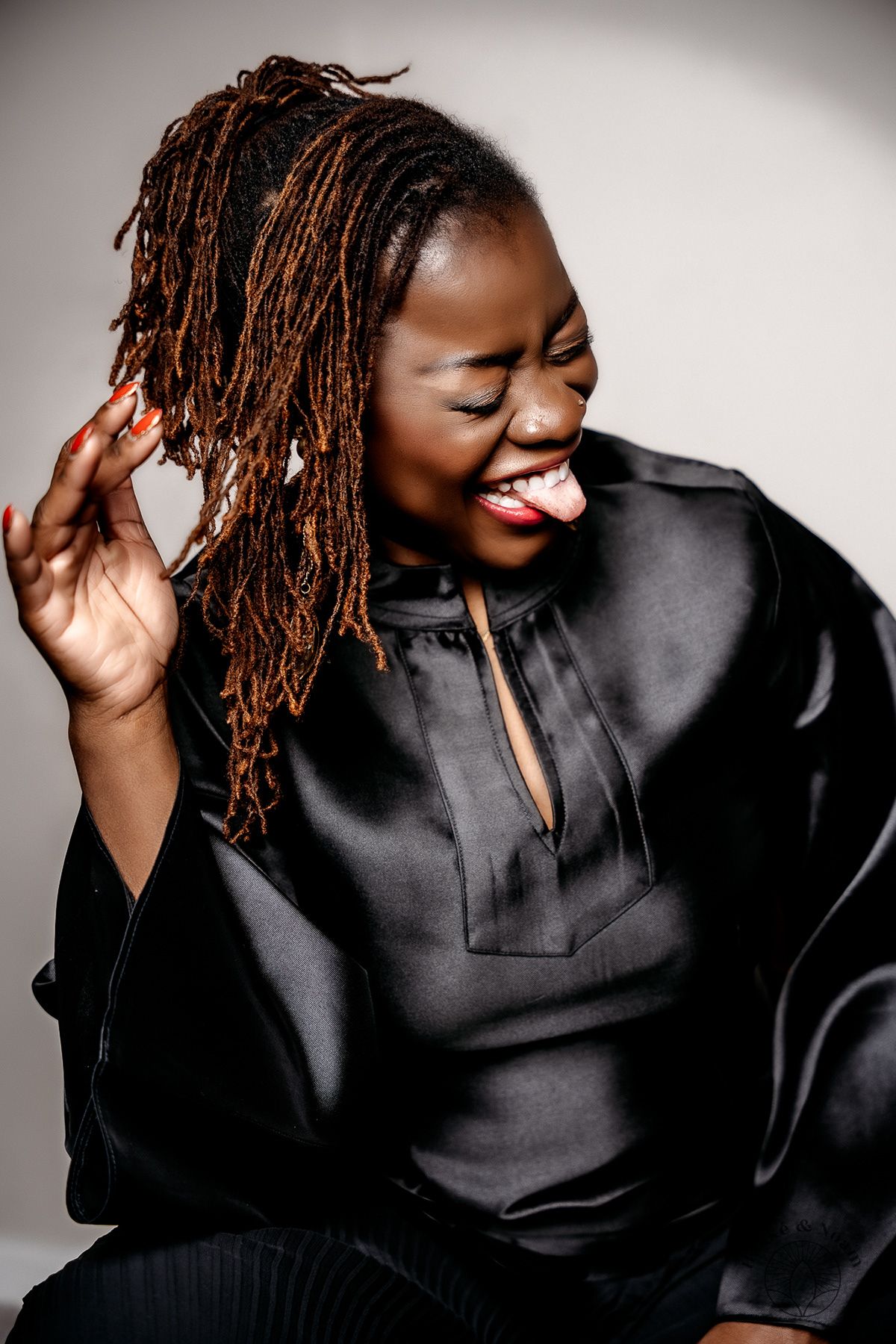Portrait of someone in black satin clothing laughing with dreadlocks against a light background.