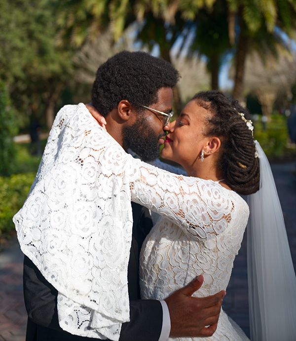 A couple shares a romantic kiss in white lace wedding attire with palm trees visible in the background.