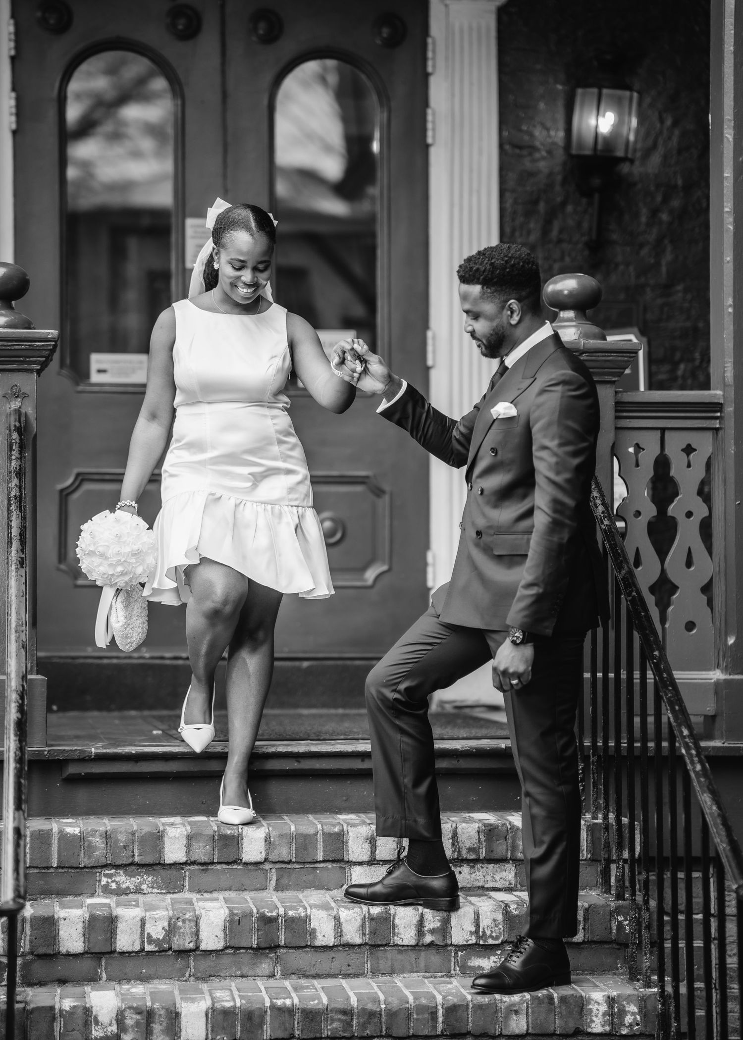 A black and white wedding photo shows a couple sharing a joyful moment on brick stairs outside a historic building.
