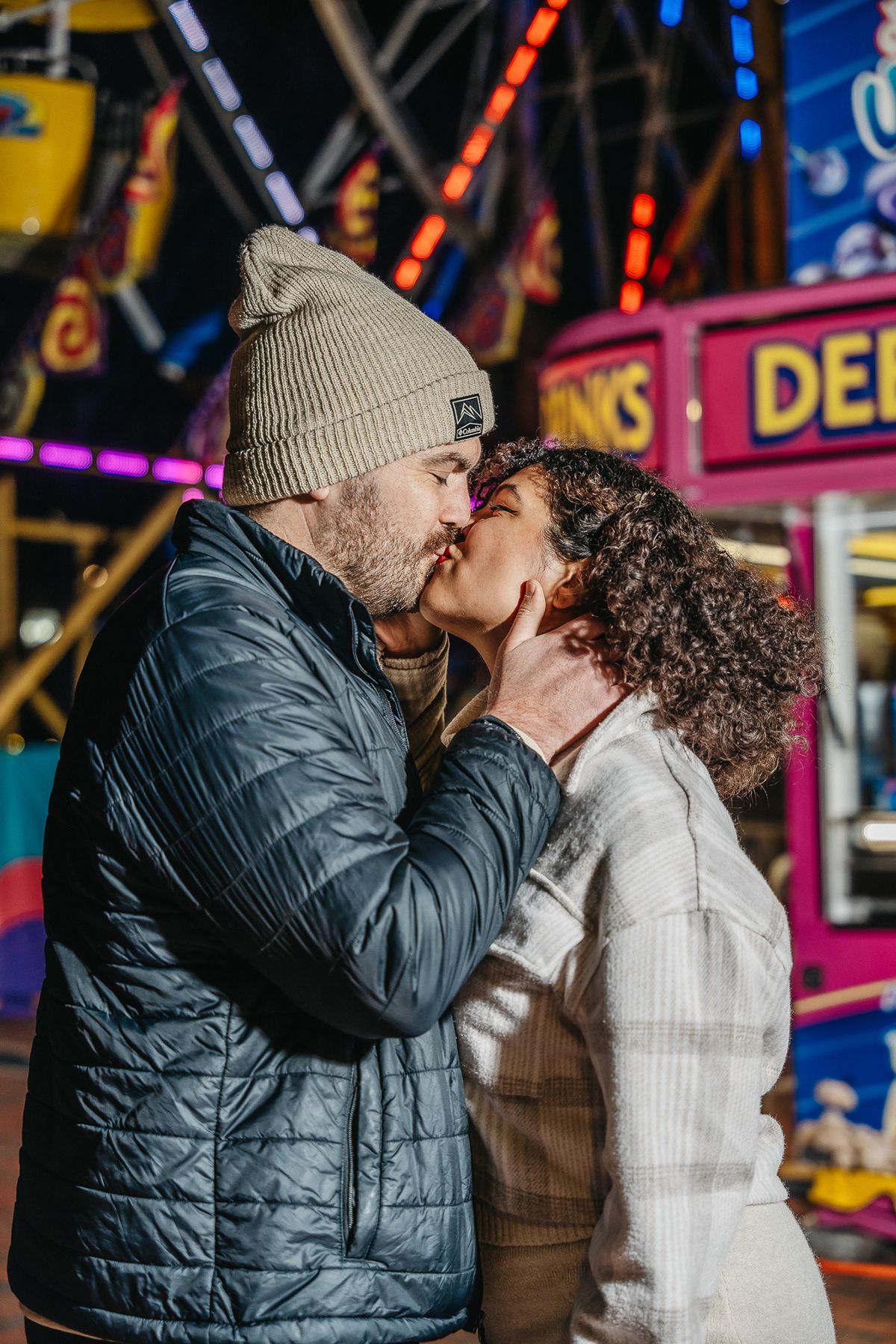 Romantic moment between a couple at a carnival with neon lights and signs in the background.