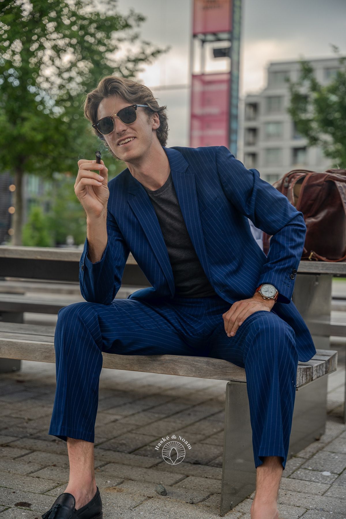 Stylish person in pinstriped navy suit relaxing on urban bench with sunglasses.