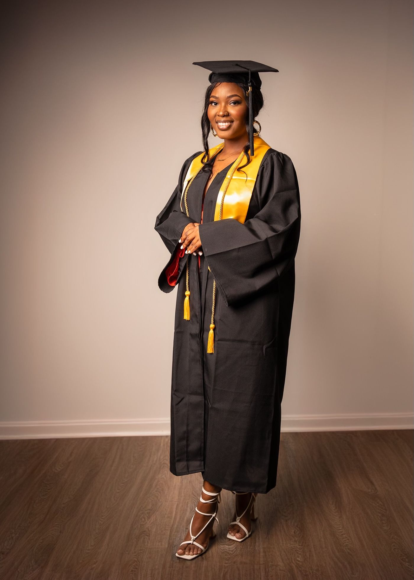 Graduate poses in black cap and gown with yellow stole and silver heels.