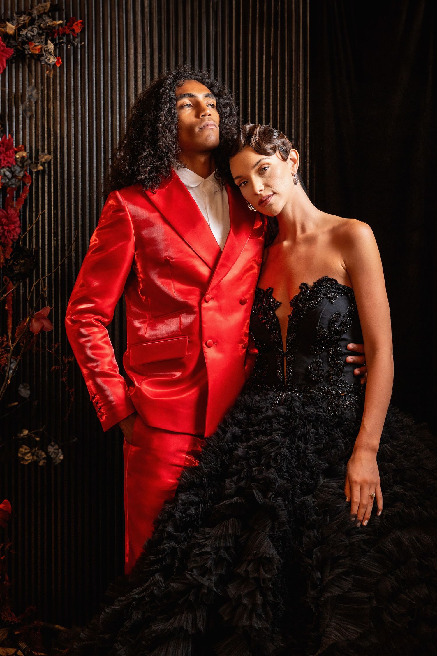 A stylish couple poses against a dark wall, contrasting a red suit with a black evening gown.