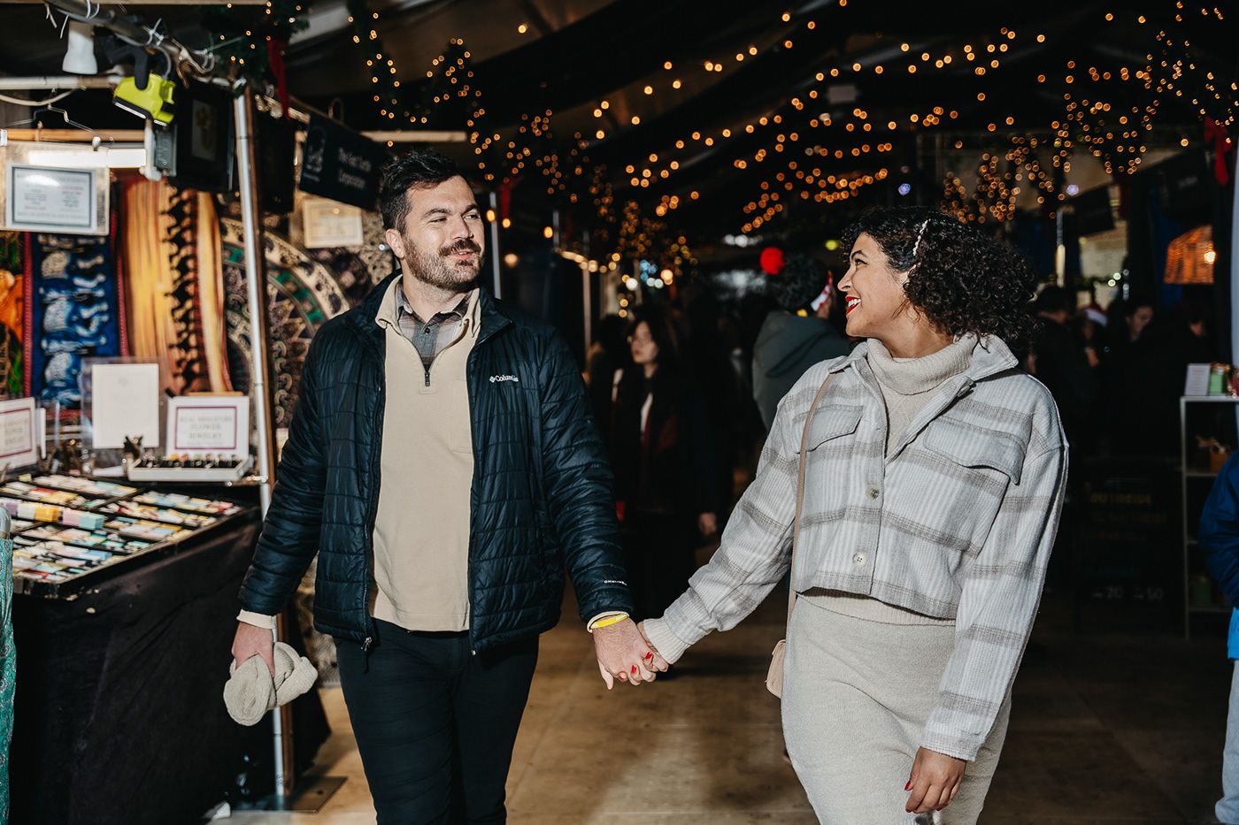 A couple walks hand in hand through an indoor market with twinkling lights overhead.