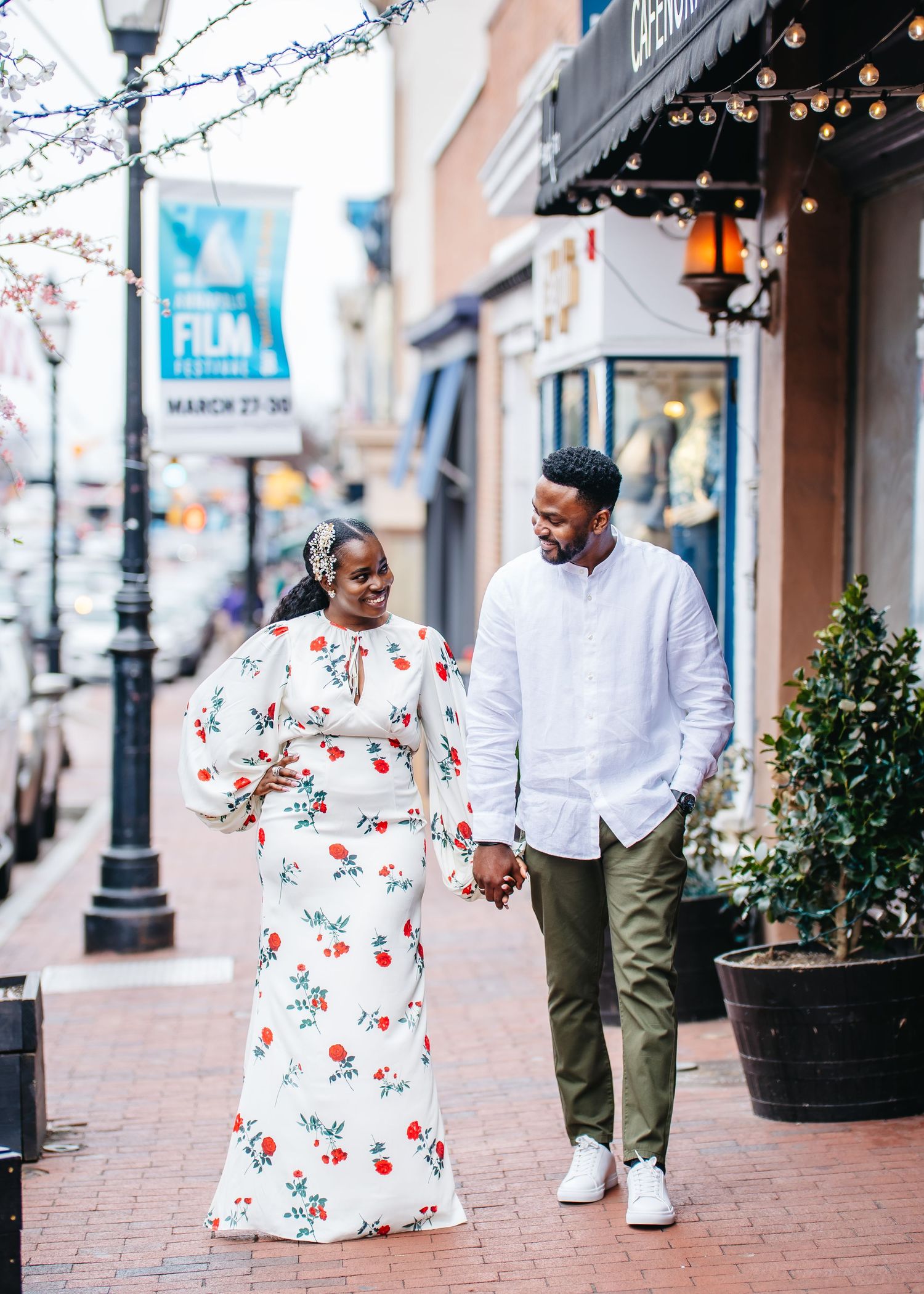 A couple walks together holding hands on a brick sidewalk in front of shops decorated with twinkling lights.