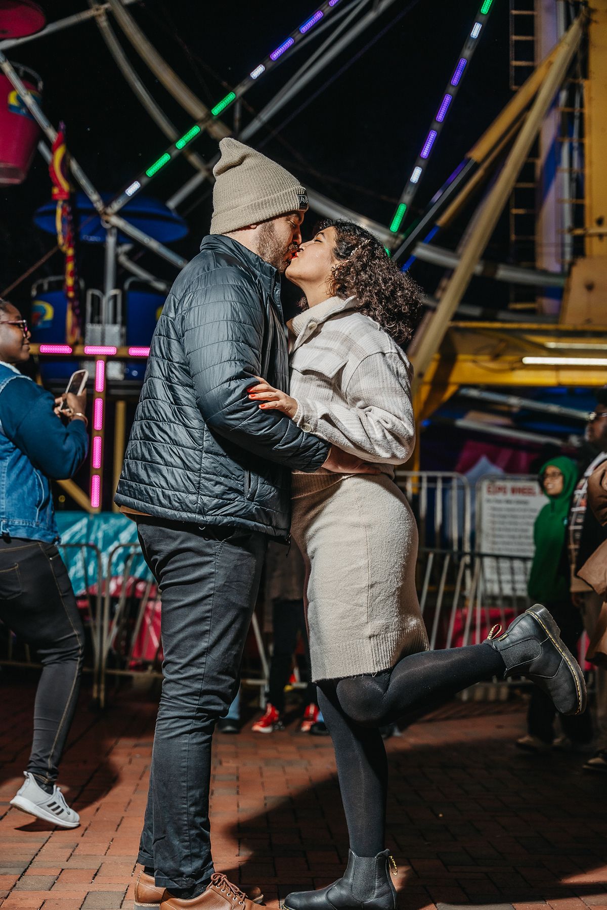 Couple embracing at a carnival with colorful lights and rides in the background.