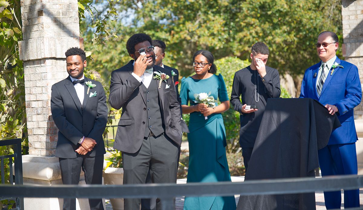 Wedding party in formal attire stands on outdoor balcony during ceremony.