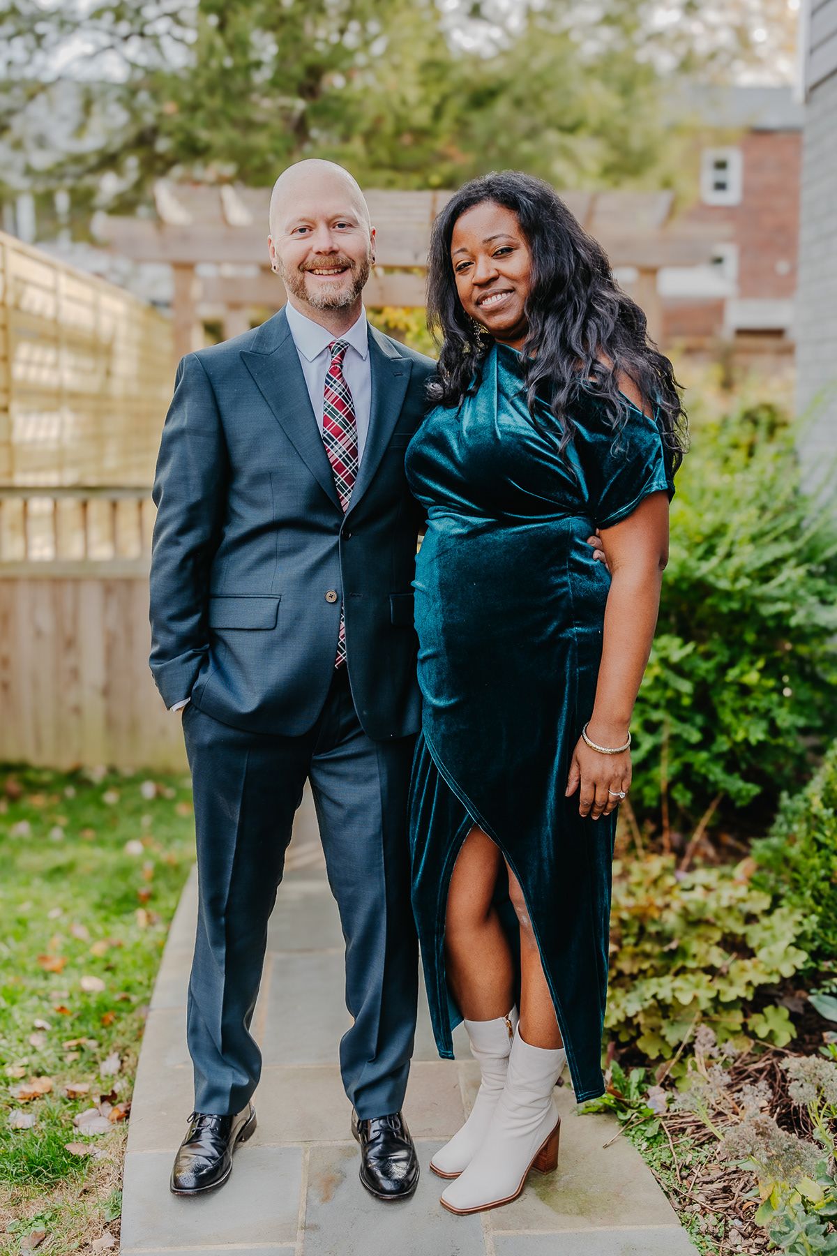 A couple stands on a garden path wearing elegant formal wear in green velvet and grey.