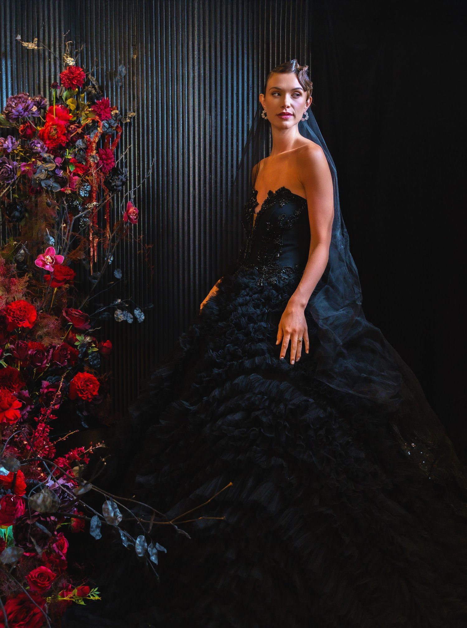 A dramatic bridal portrait with a black ruffled gown beside deep red floral arrangements.
