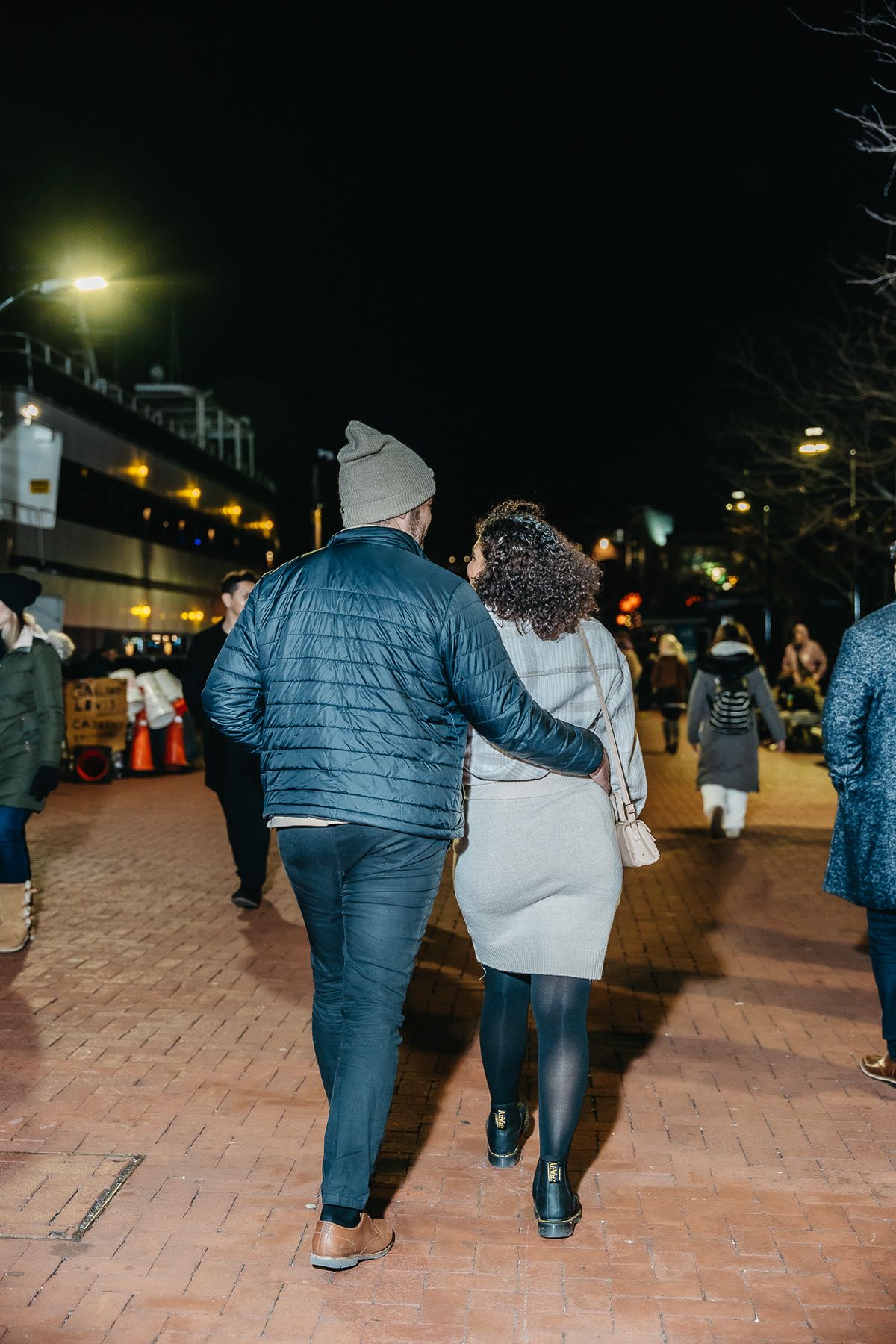 Couple walking together at night on a brick pathway with city lights in the background.