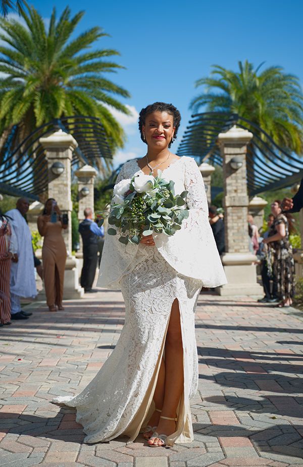Bride in elegant white lace gown holding bouquet poses at tropical wedding venue.