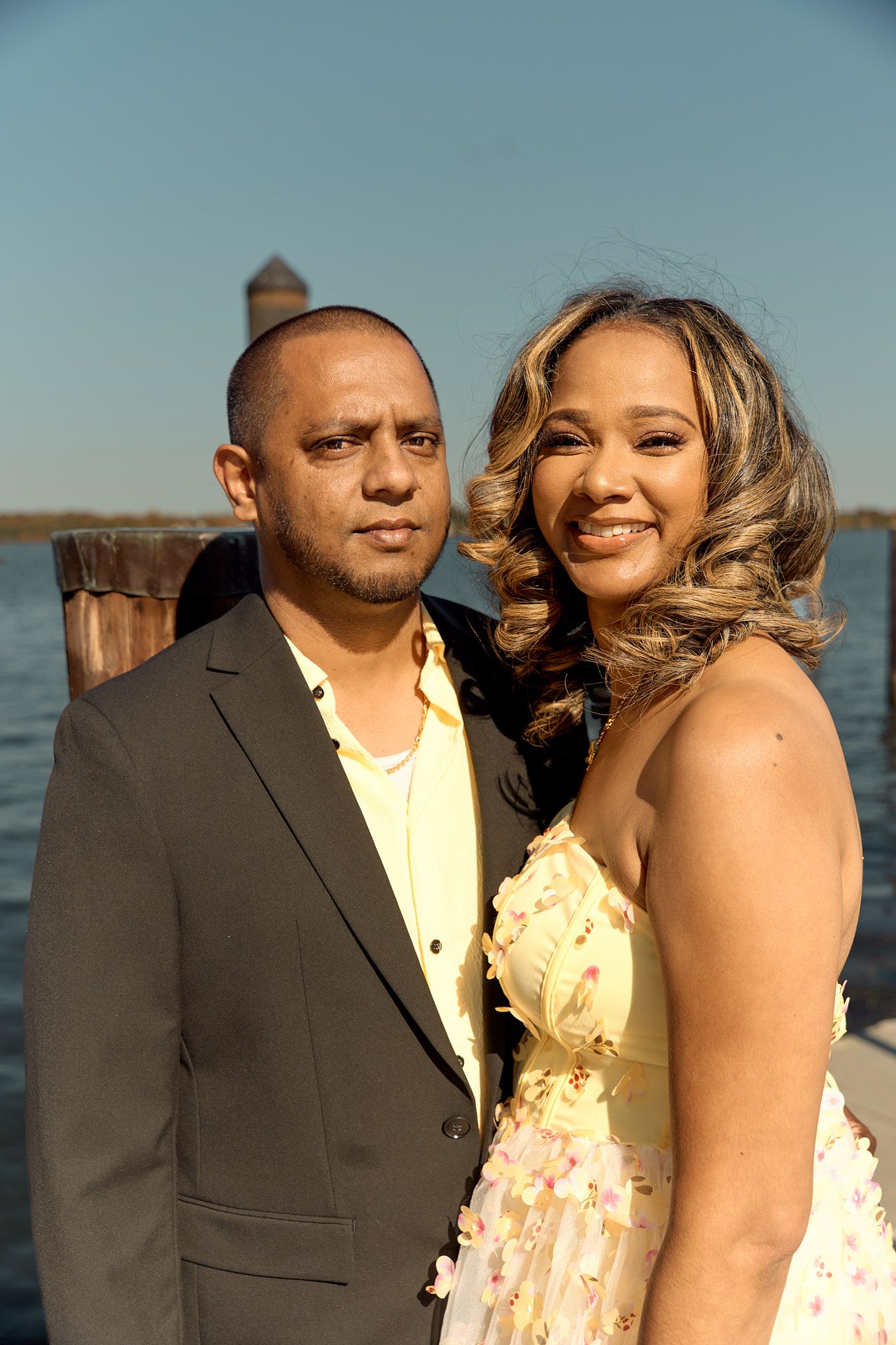 A couple in formal wear poses together on a dock with water and blue sky behind them.