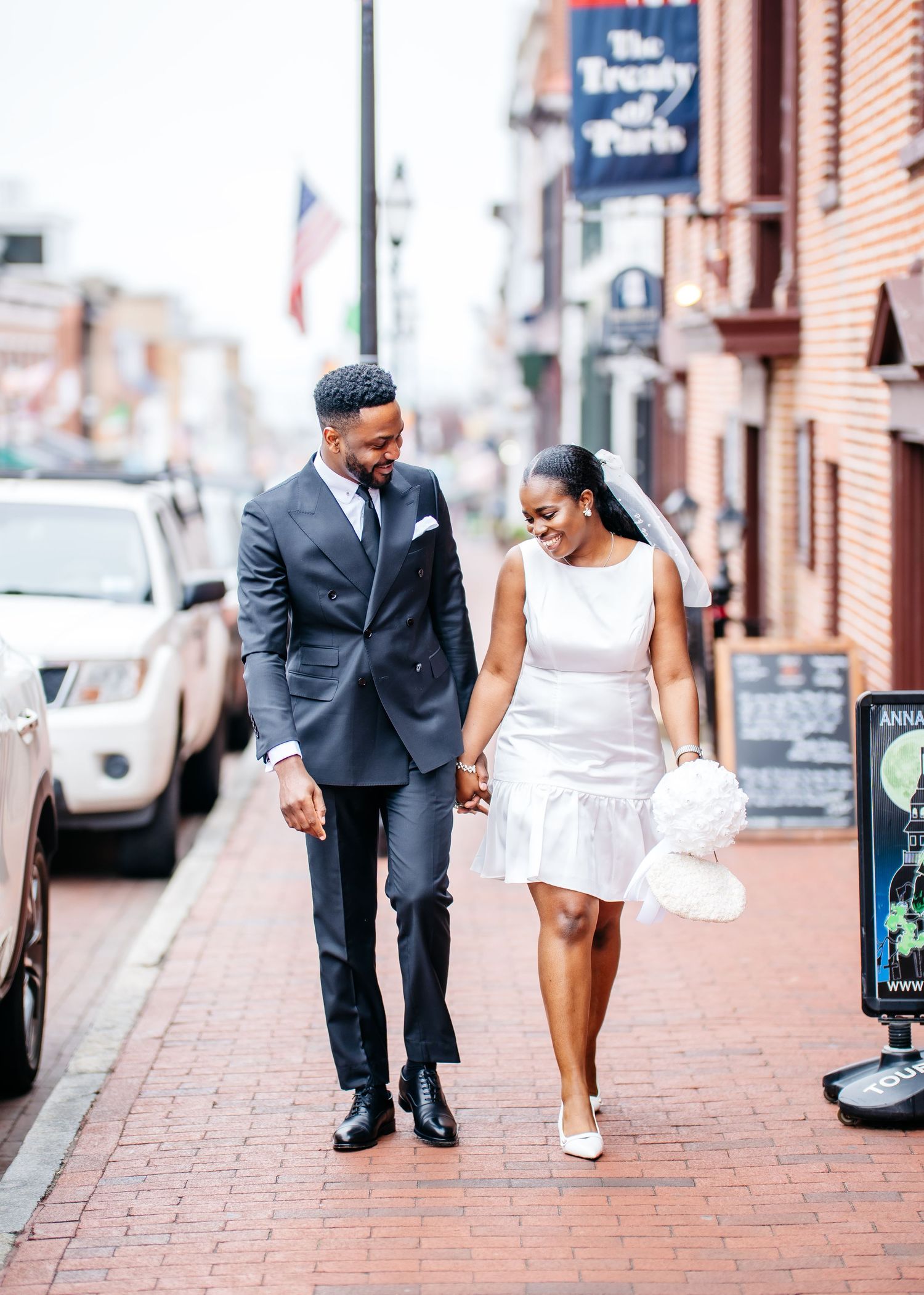 A newlywed couple walks hand in hand down a brick sidewalk in a historic district wearing formal wedding attire.