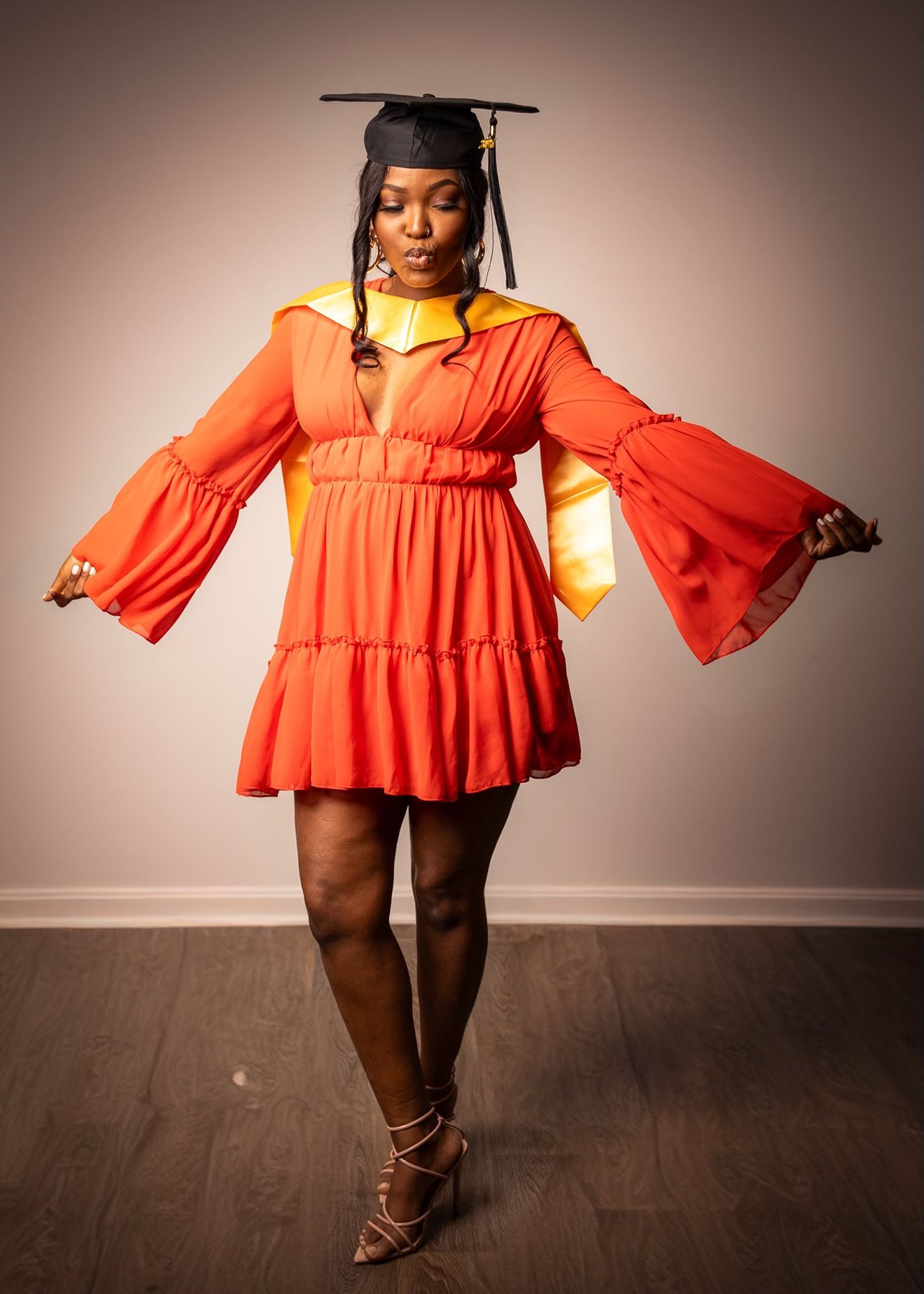 Graduate in orange bell sleeve mini dress and cap strikes a playful pose on hardwood floor.