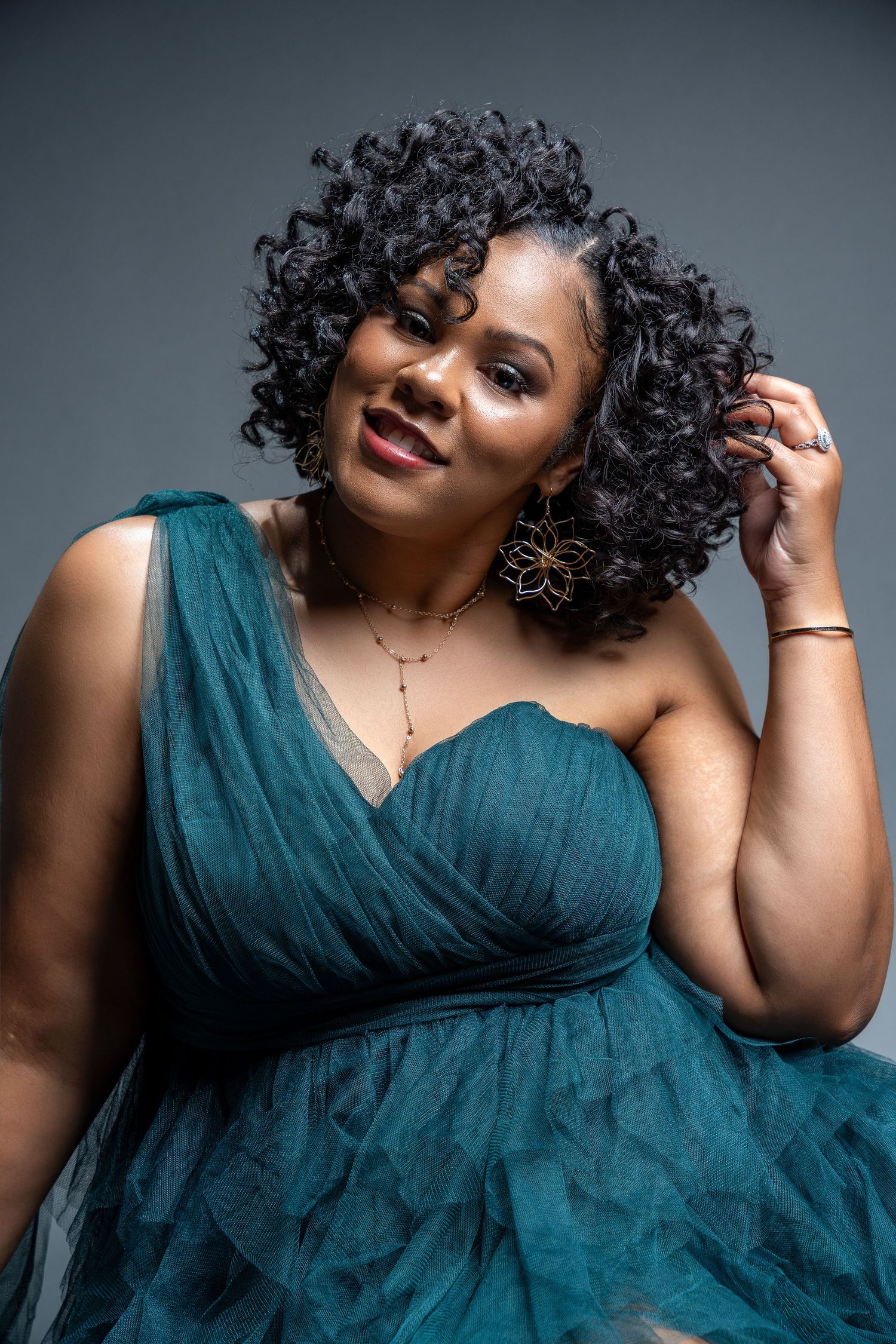 Elegant fashion portrait featuring a teal one-shoulder evening gown and natural curly hairstyle against gray background.