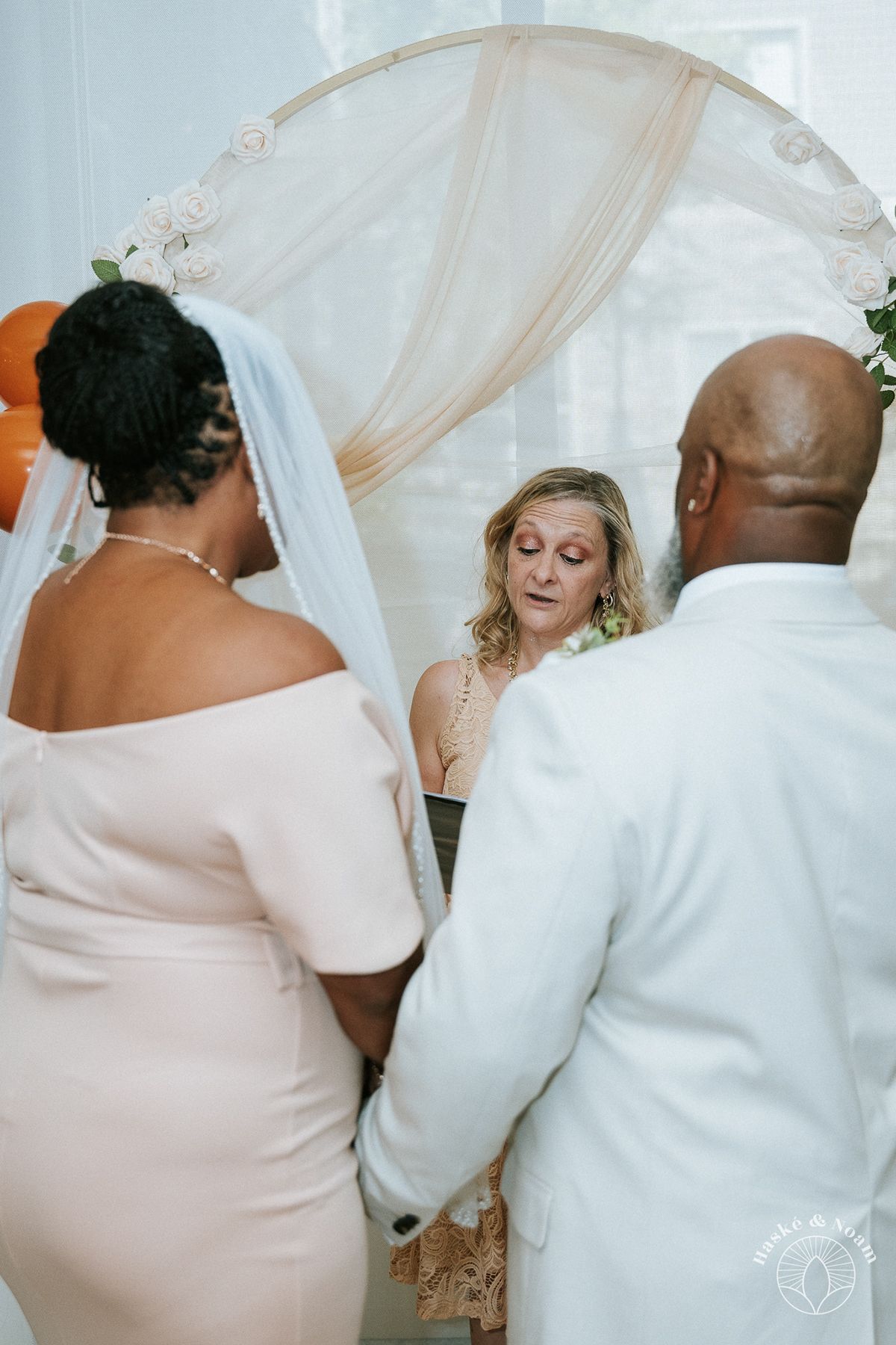 Wedding ceremony officiant in black dress stands under arch with couple during intimate ceremony.