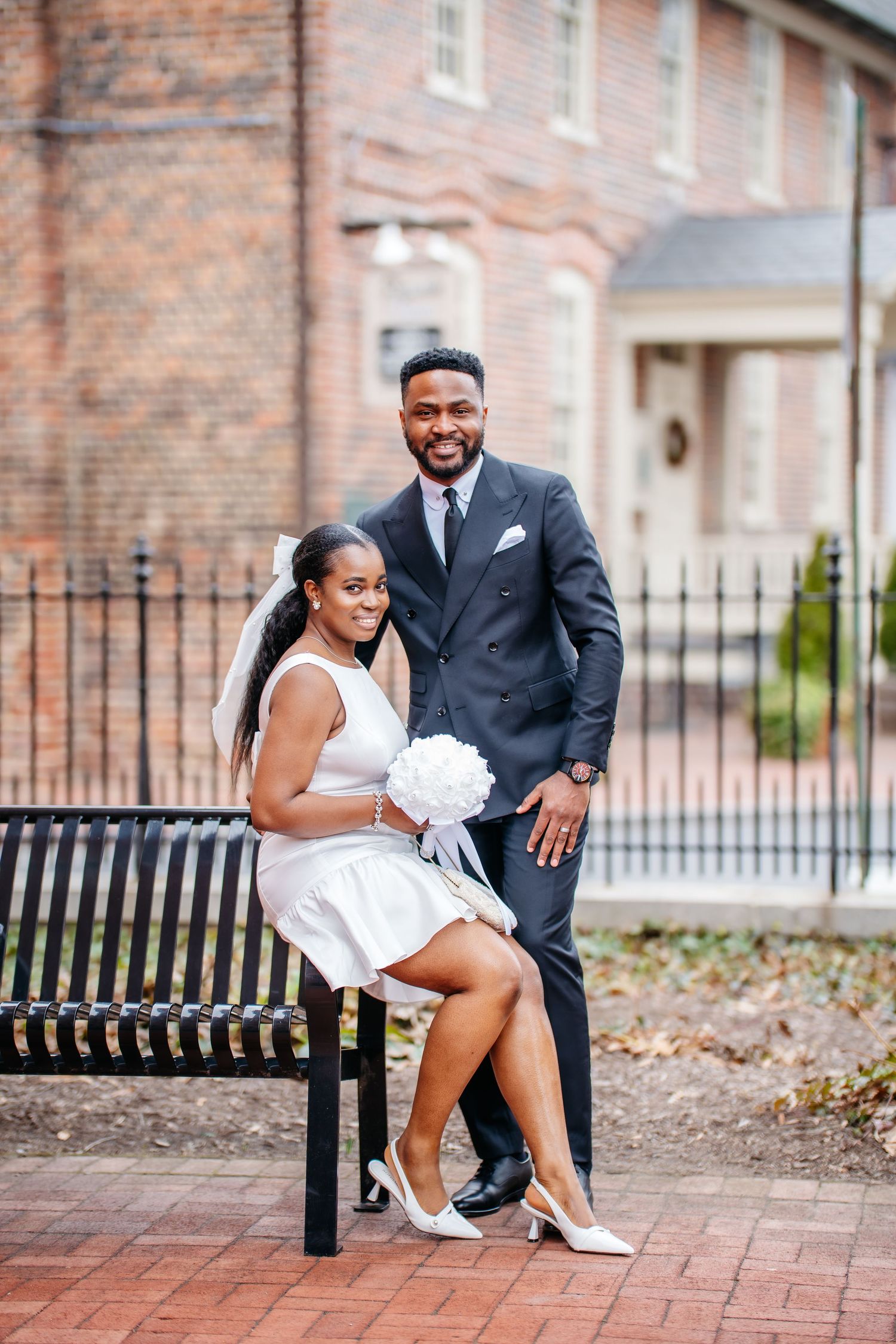 Newly married couple in formal attire sitting on a bench by a brick building with a white floral bouquet.